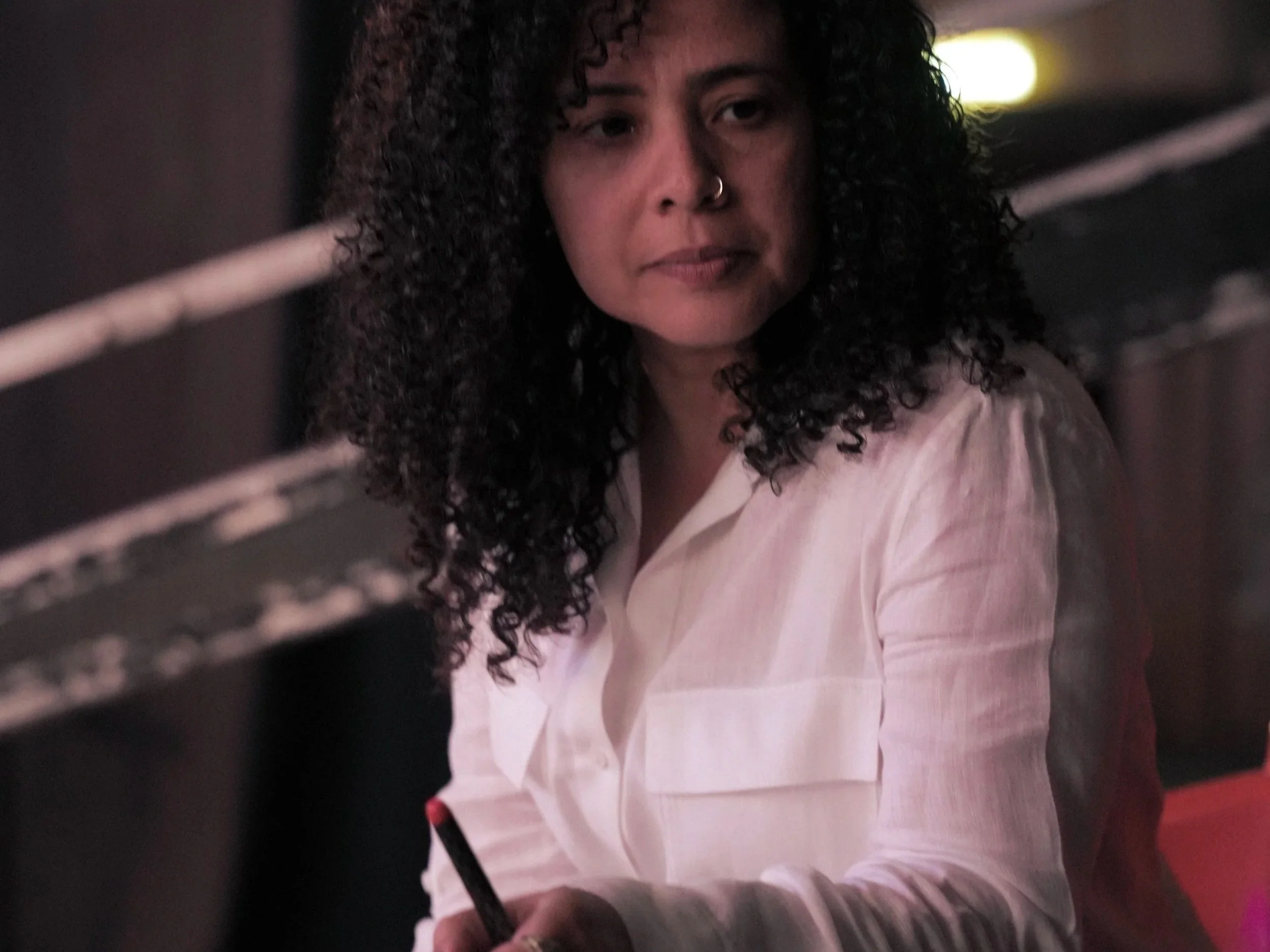A woman with dark curly hair, wearing a nose ring and a light-colored shirt, looking at the camera with a neutral expression. The photo appears to be taken through metal bars or mesh.