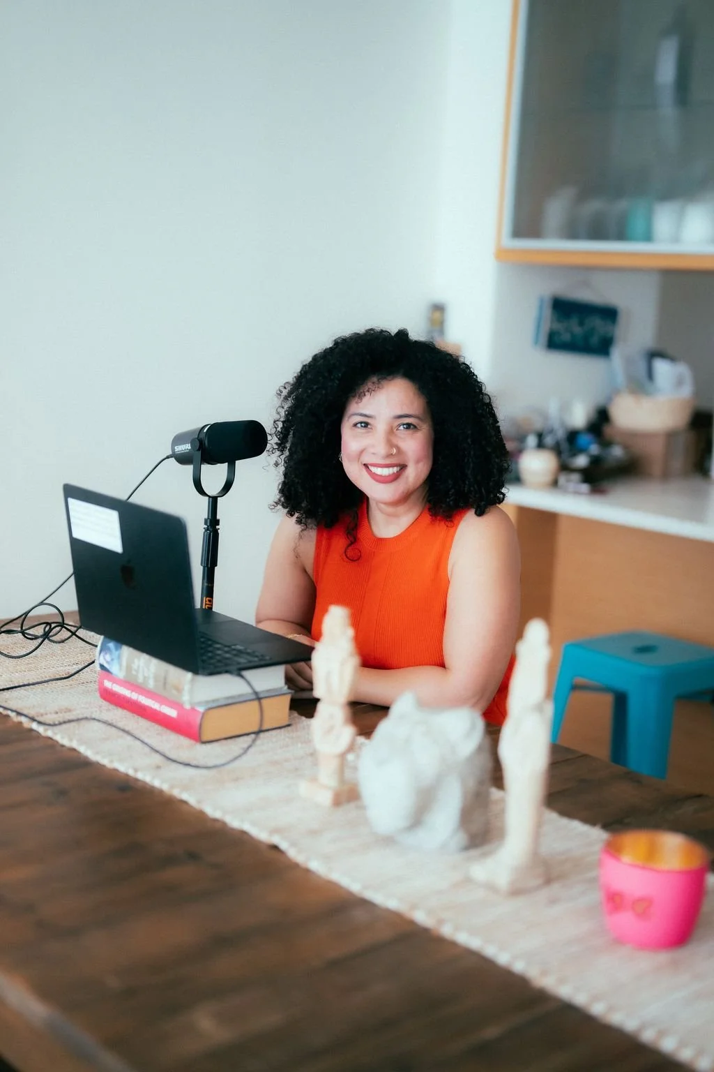A woman with curly black hair and a bright orange sleeveless top, smiling and sitting at a dining table with a laptop, microphone, and decorative figurines, in a cozy kitchen or dining area.
