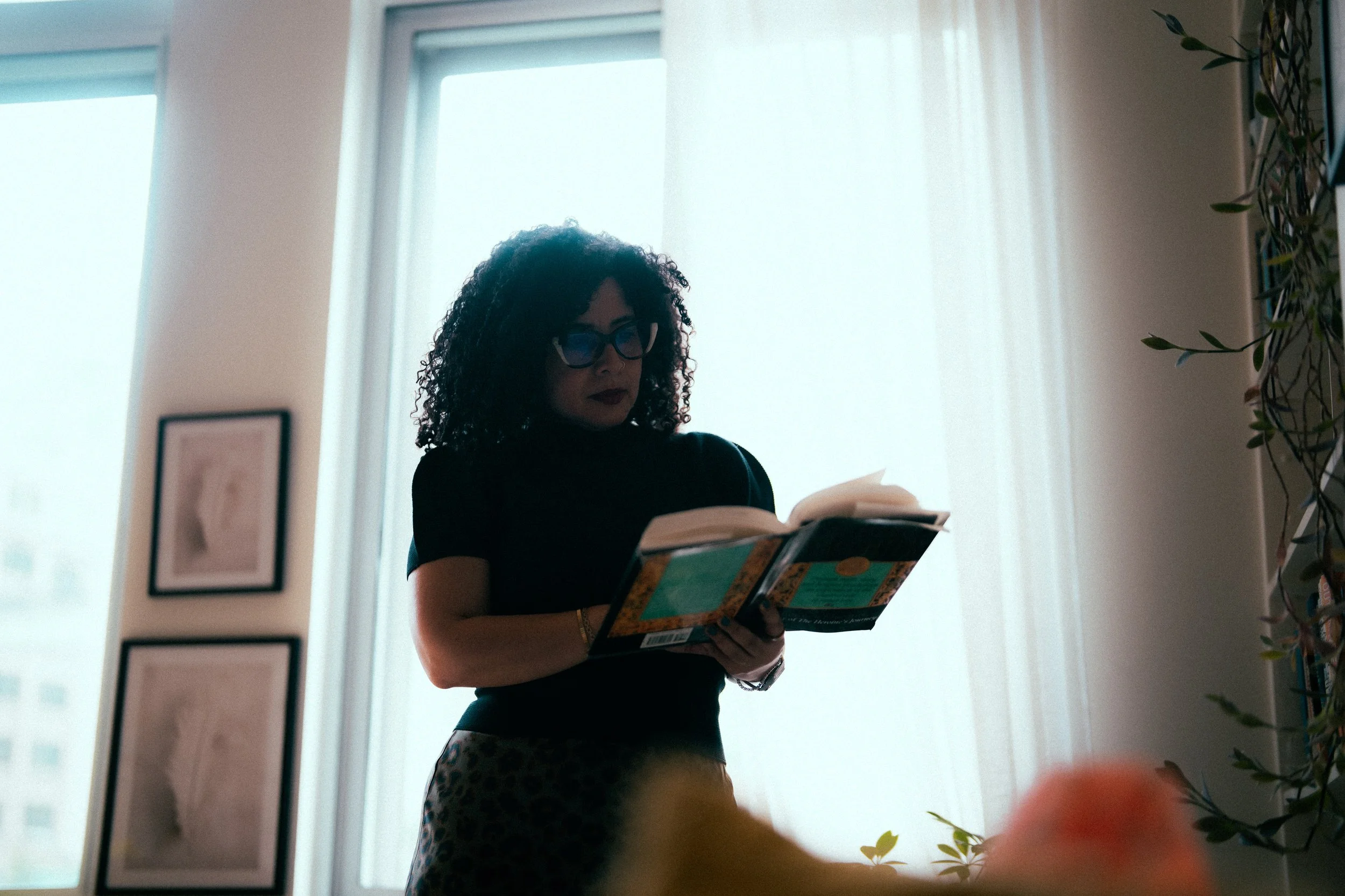 A woman with curly hair and glasses reading a book in front of large windows with light coming through, and some framed artwork on the wall.
