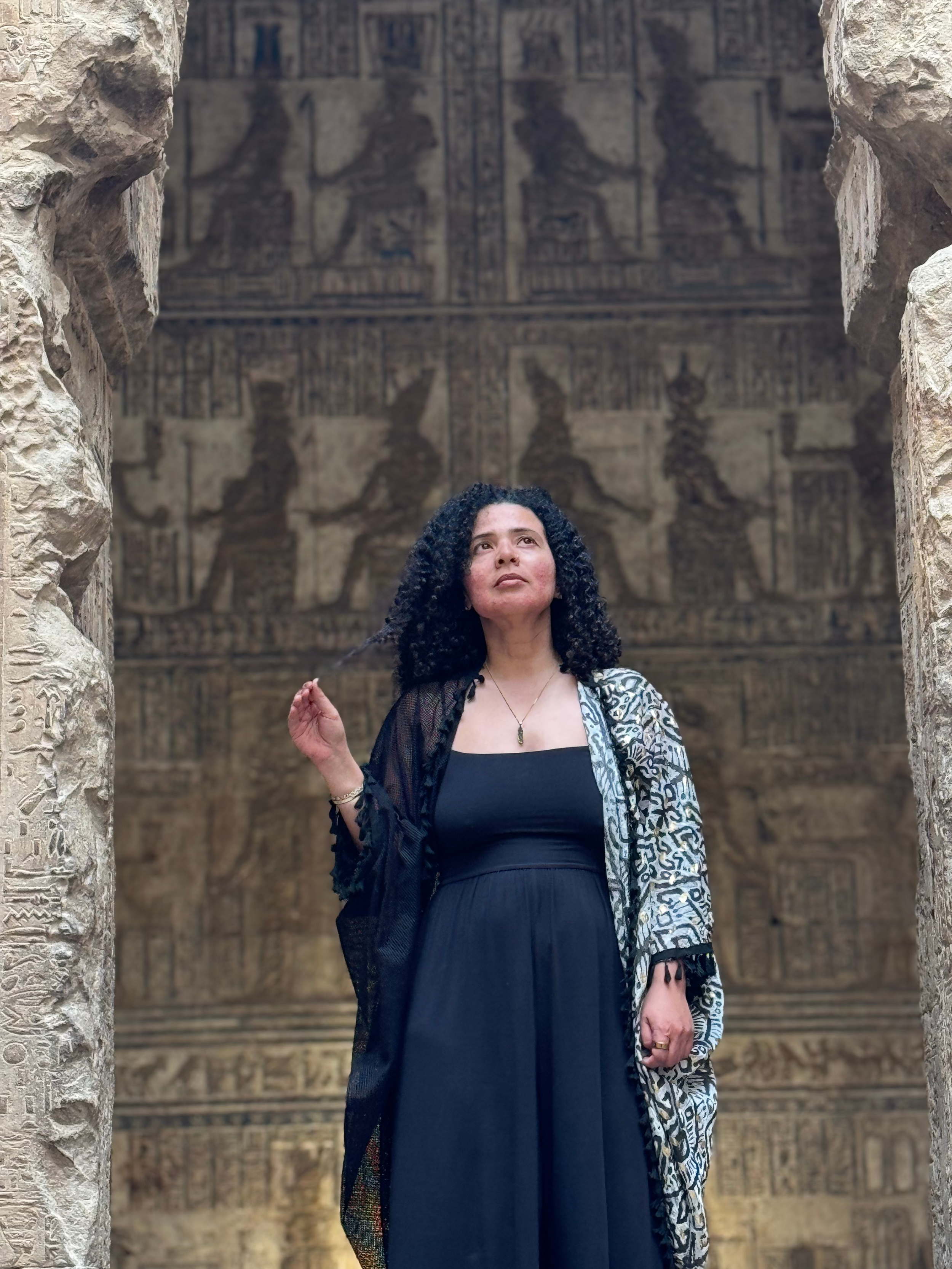 A woman with curly black hair standing in front of ancient Egyptian hieroglyphic carvings and wall paintings at an archaeological site.