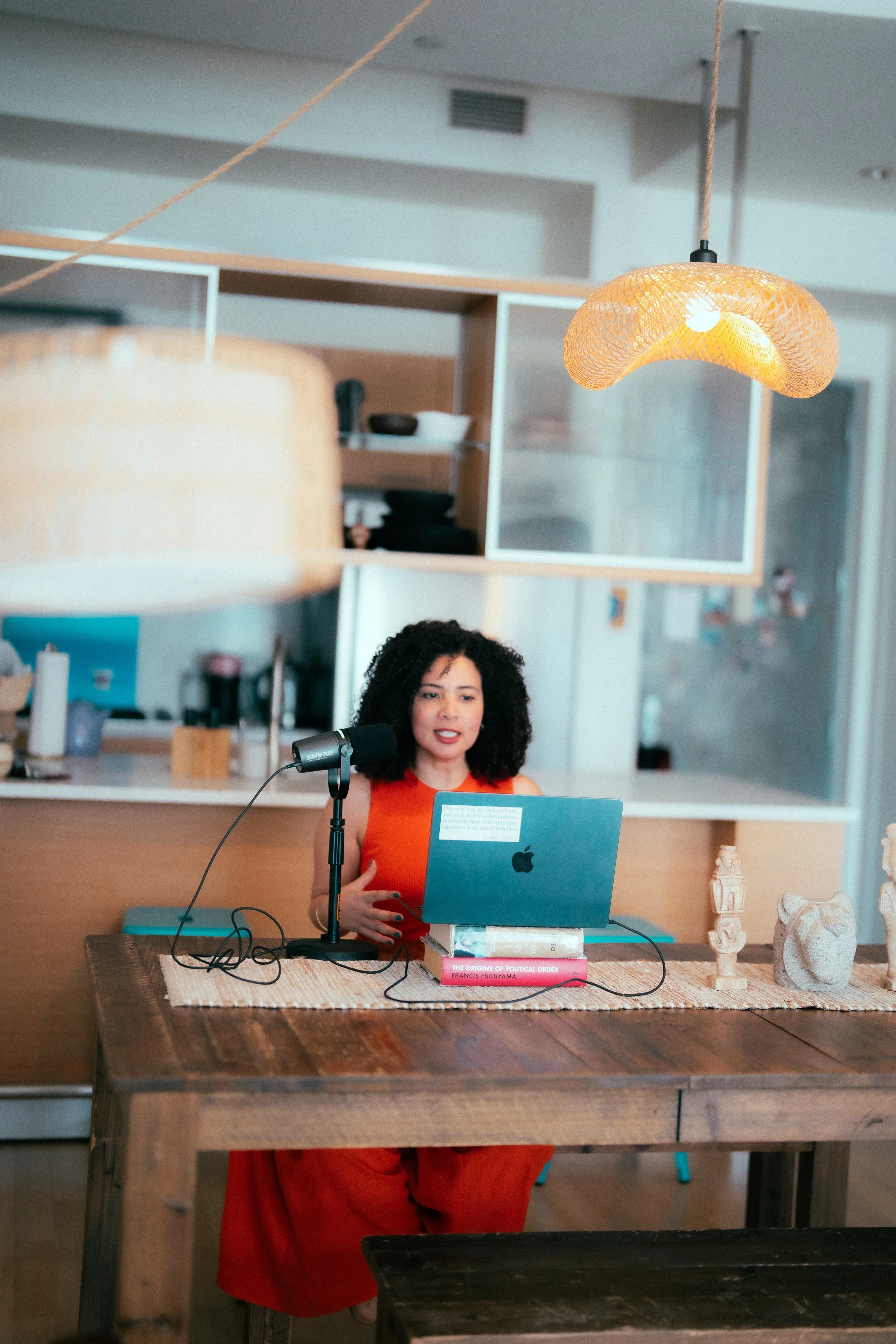 A woman with curly black hair wearing an orange dress is sitting at a wooden table with a laptop, books, and decorative objects. She is speaking into a microphone in a cozy room with shelves, art, and warm lighting.