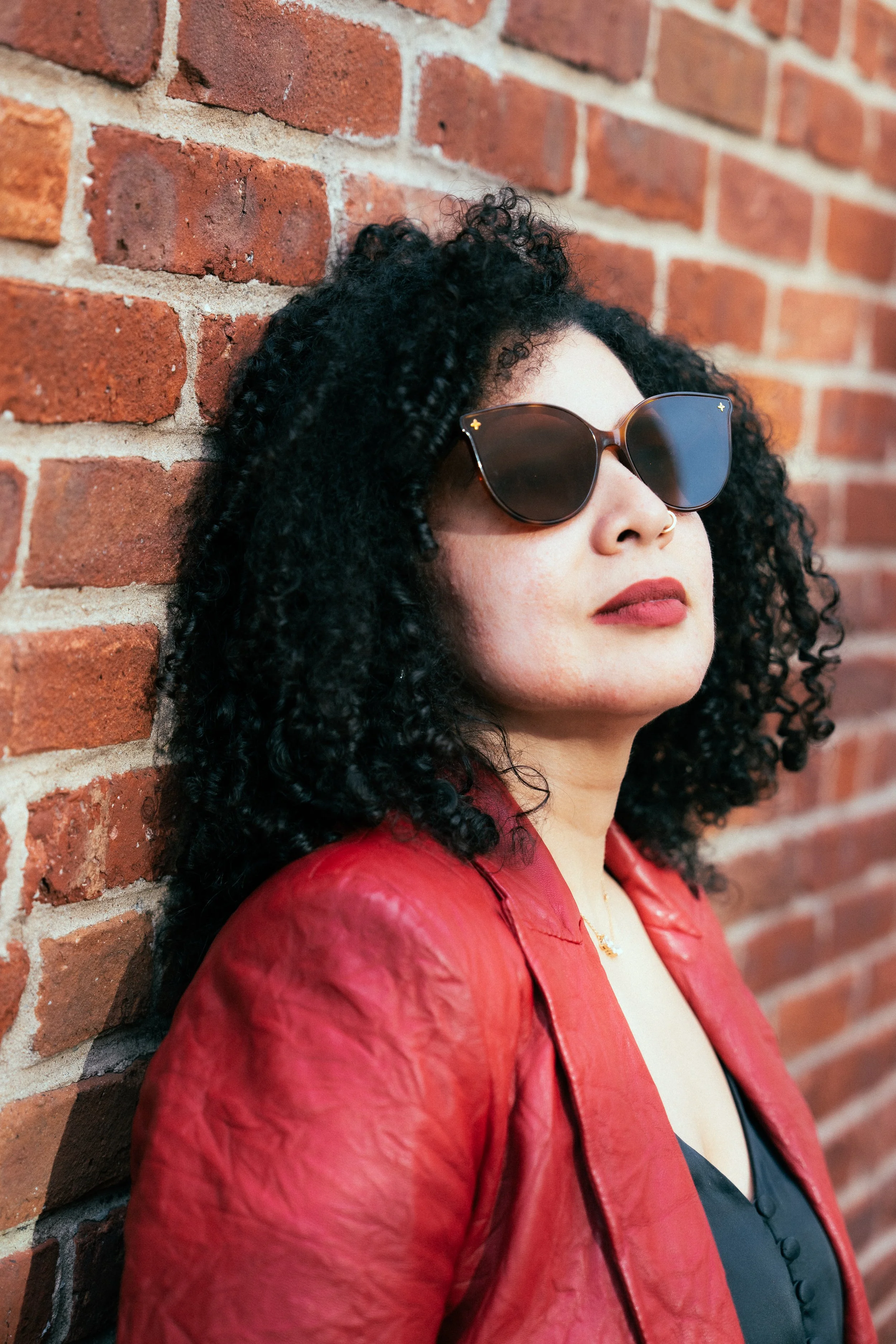 A woman with curly black hair wearing sunglasses and a red leather jacket, leaning against a brick wall.