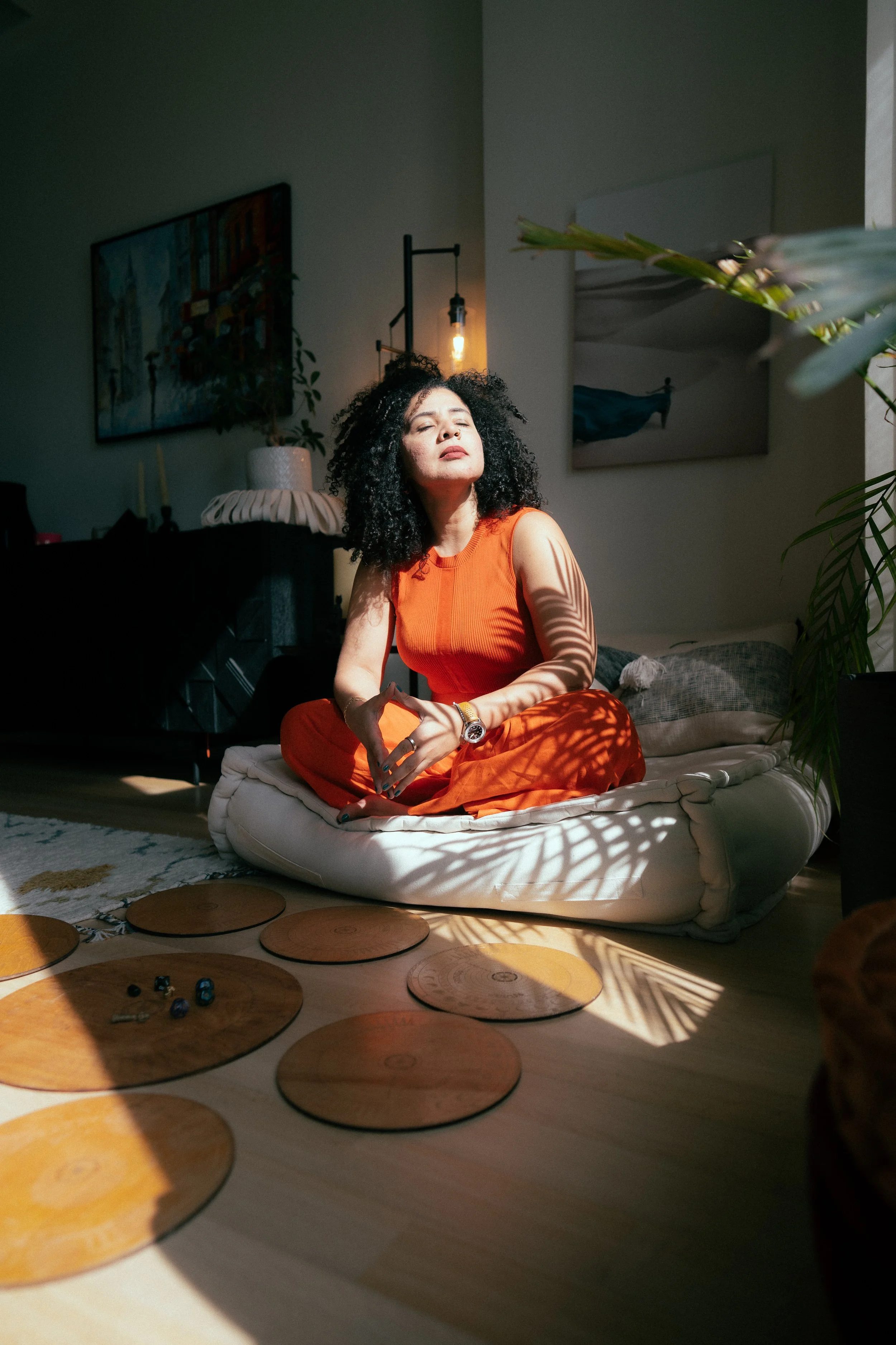 A woman with curly hair and wearing an orange sleeveless top and pants, sitting cross-legged on a plush cushion in a room with warm sunlight streaming through window blinds. She's looking up with her eyes closed, surrounded by indoor plants, wall art, and a cozy interior setting.