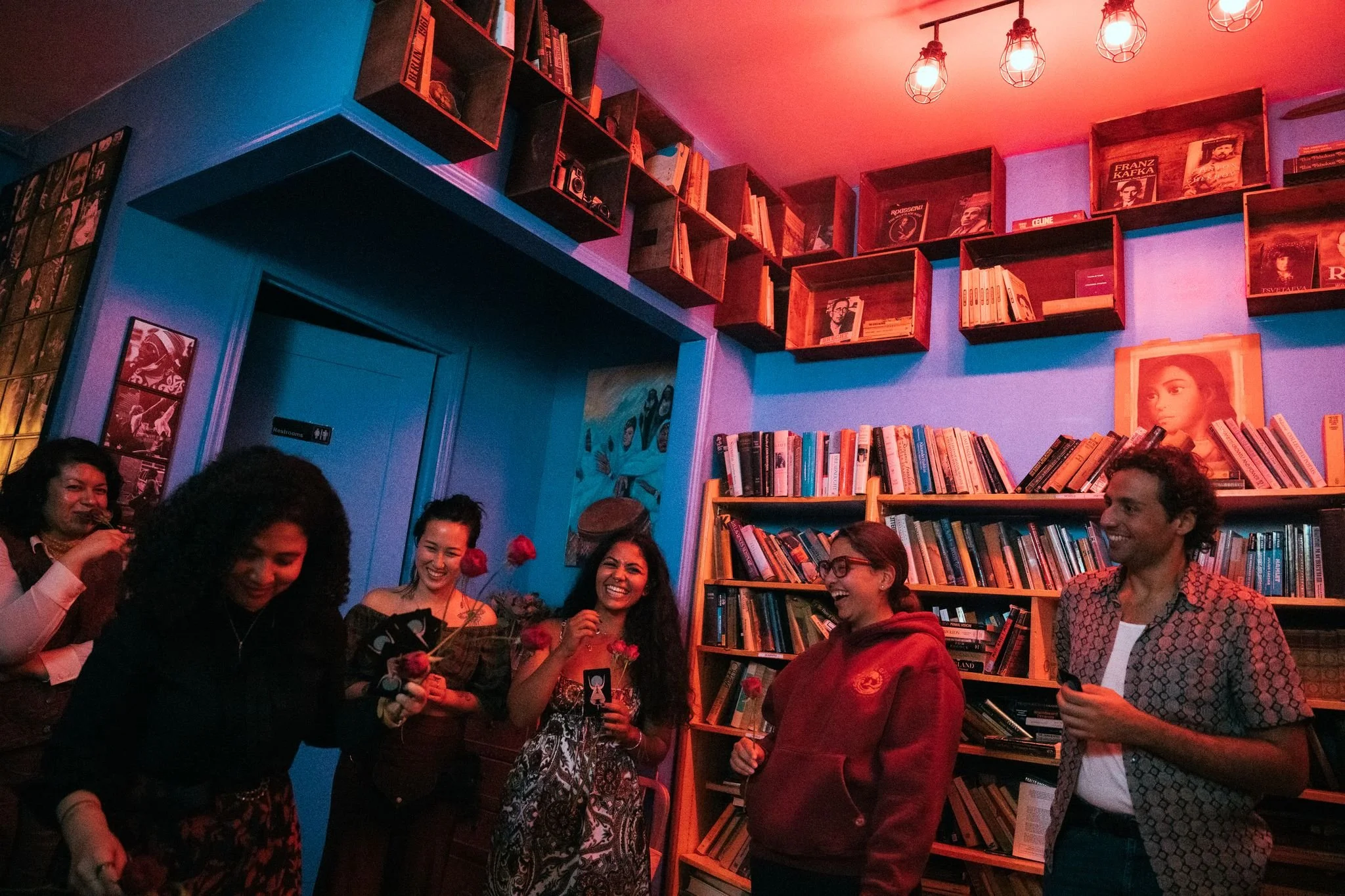 People gathered in a cozy, dimly-lit room with bookshelves and colorful lights, smiling and socializing at a party or gathering.