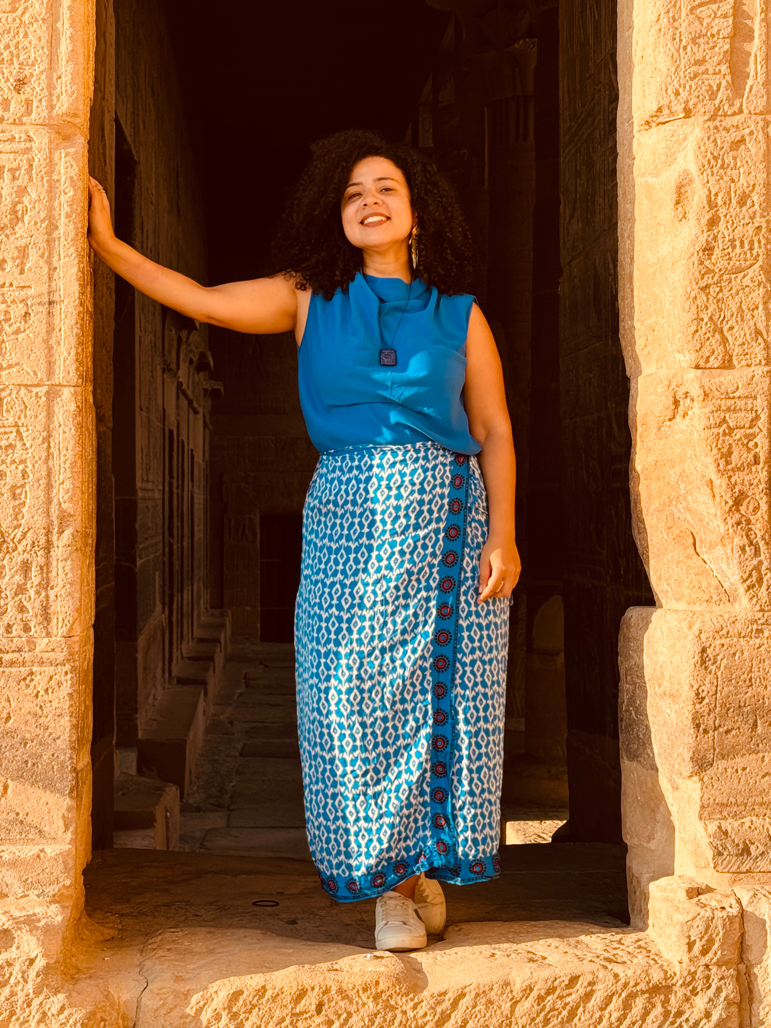 A woman with curly hair wearing a blue sleeveless top and a patterned blue and white skirt, smiling and standing in a stone doorway with her arm resting on the side, inside a historical or ancient structure.