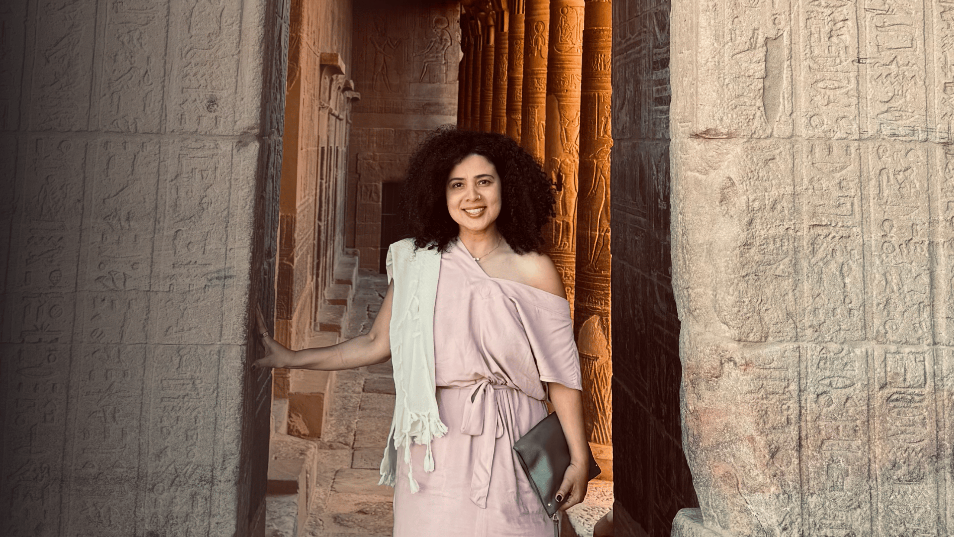 A smiling woman with curly dark hair, wearing a light pink off-shoulder dress and a cream shawl, standing amid ancient Egyptian stone walls with hieroglyphs, and orange carved columns in the background.