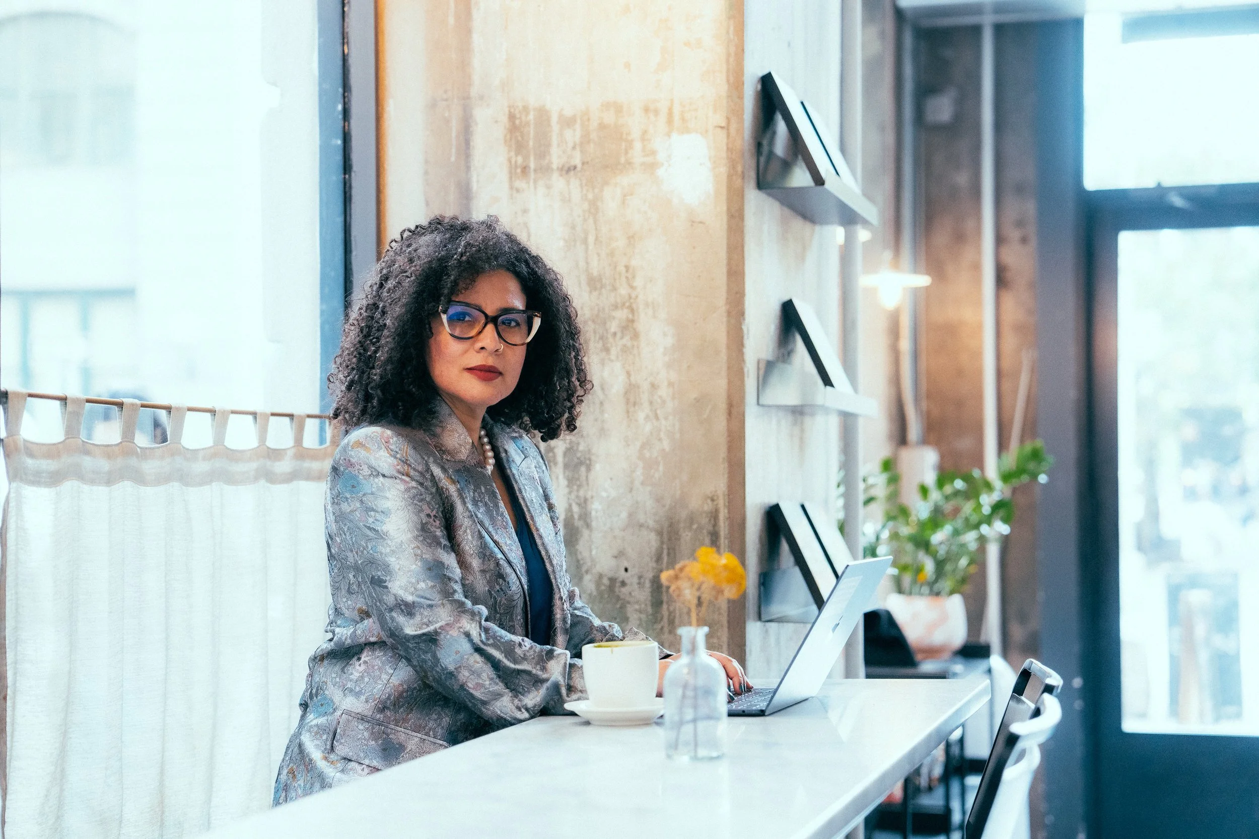 Woman with curly hair wearing glasses and a patterned blazer working on a laptop at a white counter in a modern, well-lit cafe or workspace with large windows and plants.