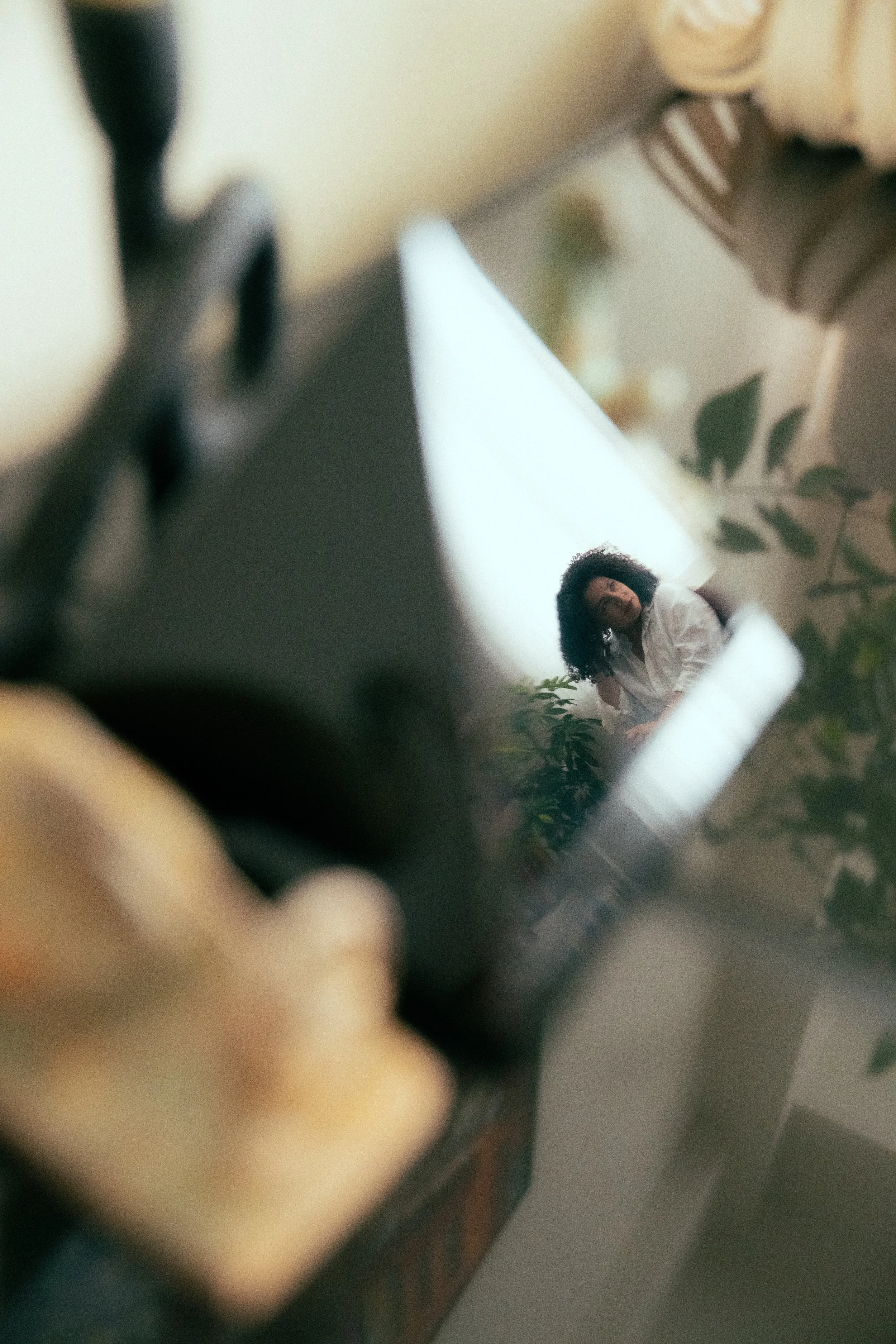 A woman with curly hair sitting next to a plant, seen through a mirror or reflective surface with some objects in the foreground.