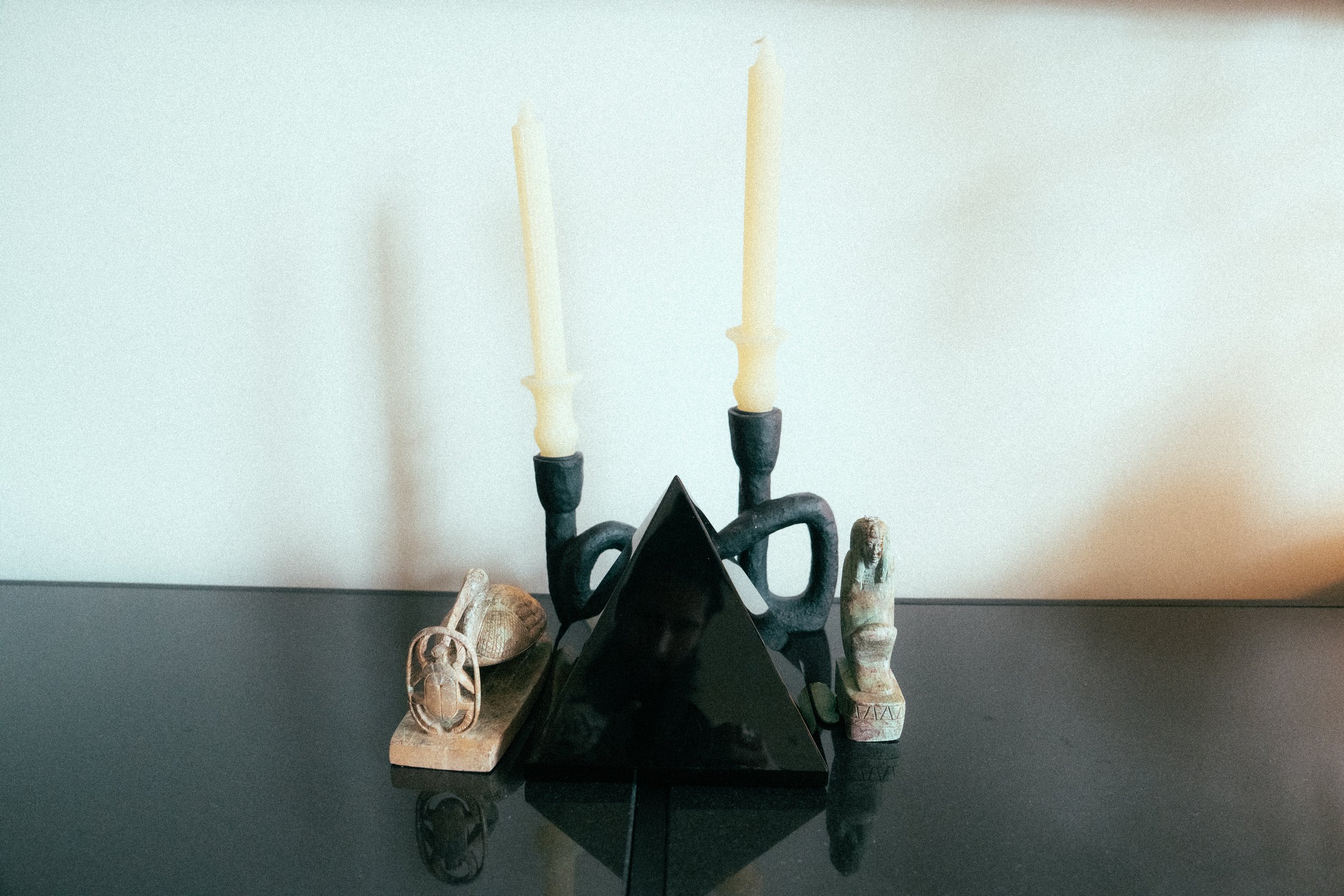 Decorative black metal candelabra with two beige candles against a white wall, surrounded by small decorative objects including carved stones and figurines, on a black surface.