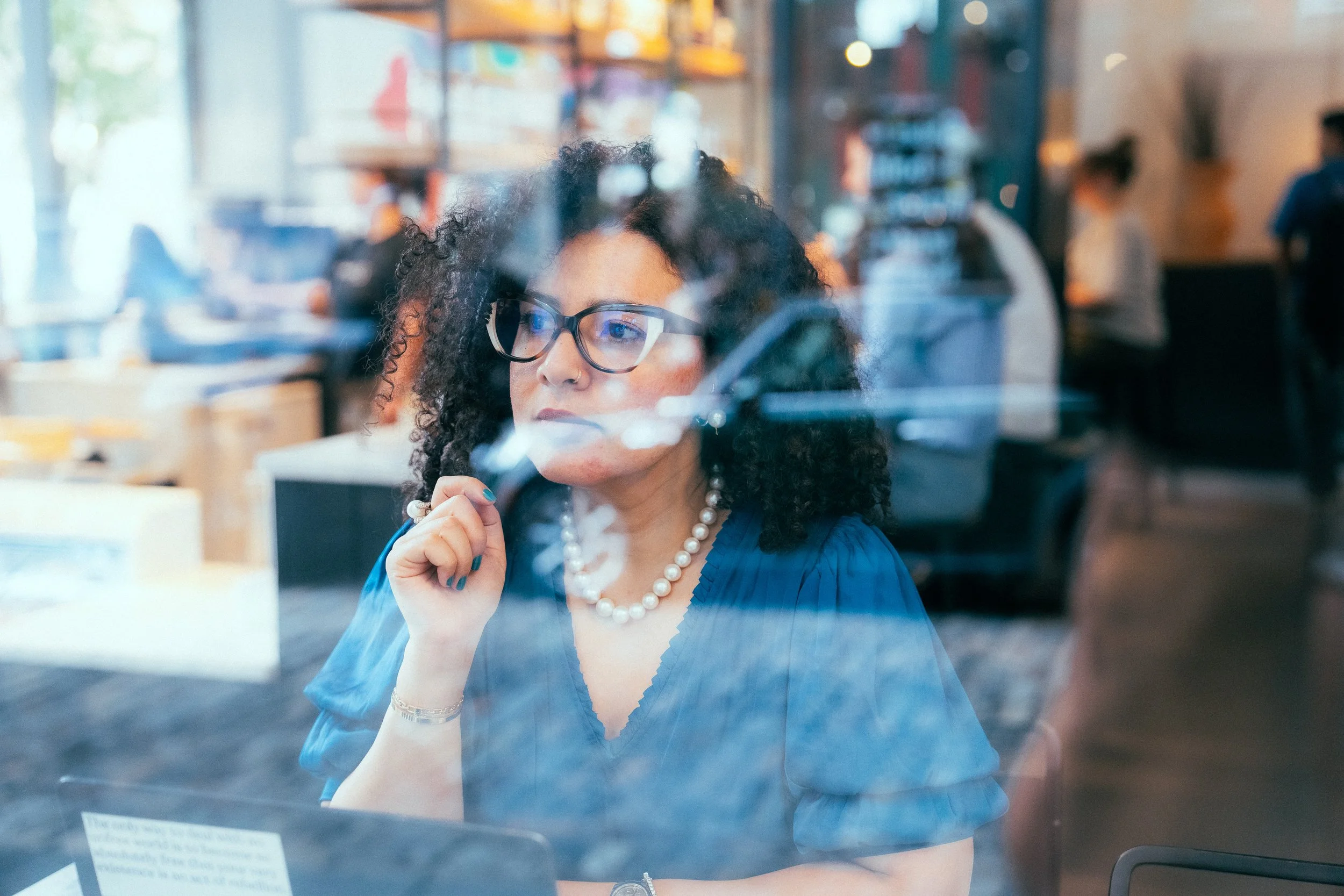 A woman with curly dark hair, wearing glasses, a pearl necklace, and a blue blouse, seen through a window in a busy café or workspace, holding a pen near her chin with a focused expression.