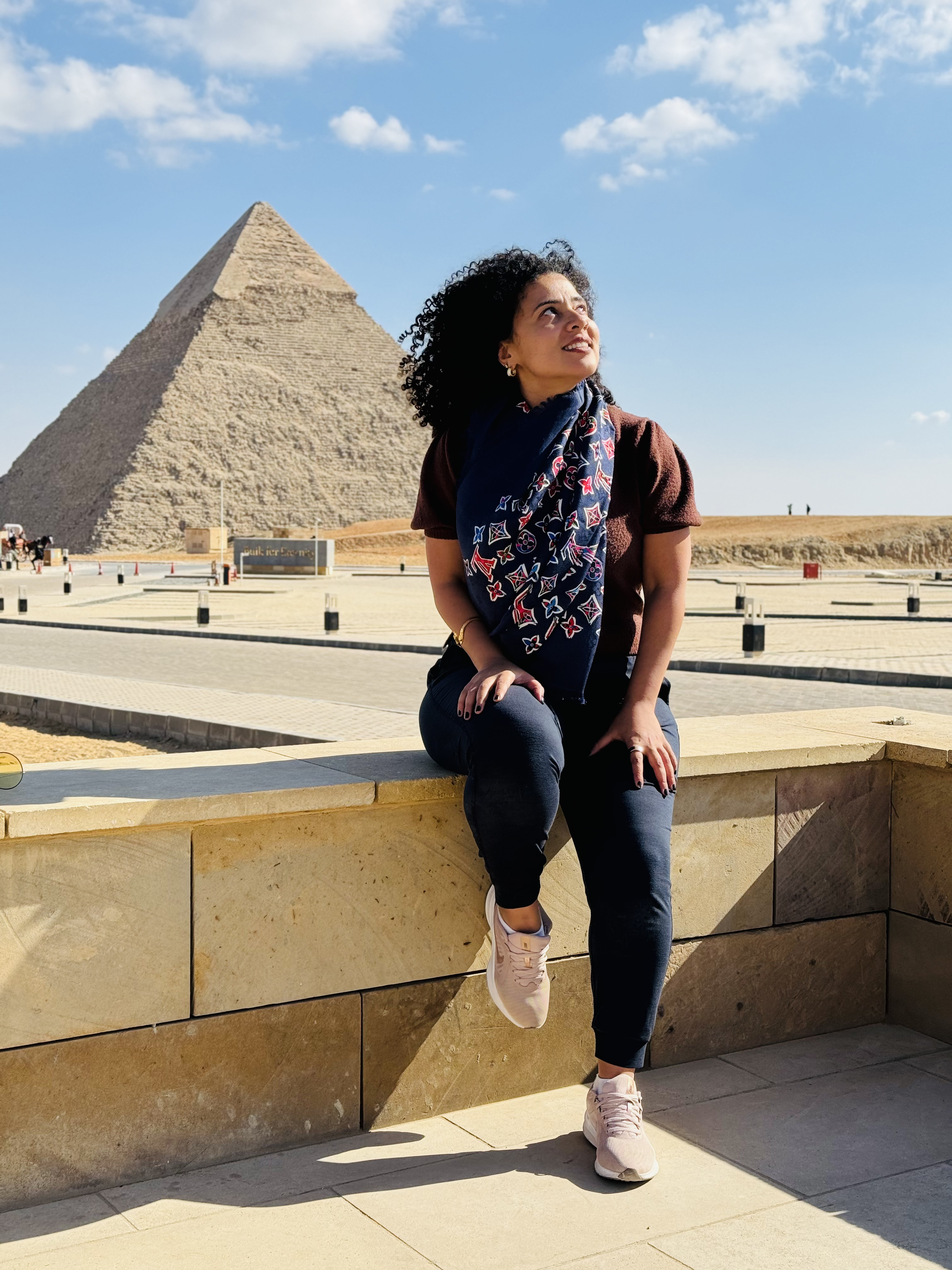 A woman with curly hair wearing a headscarf, brown top, and athletic shoes, sitting on a stone ledge in front of the pyramids of Egypt on a sunny day with scattered clouds.