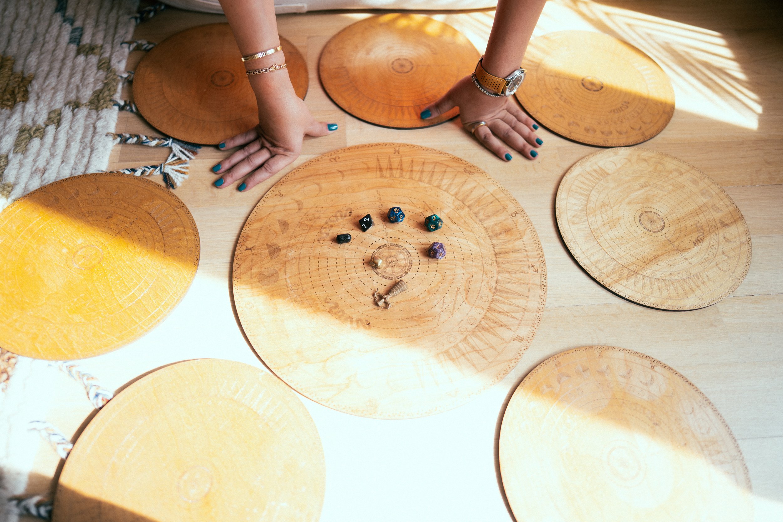 Top-down view of a person with blue painted nails and wearing gold jewelry, playing a tabletop role-playing game with round wooden game pieces and four dice on circular game boards.