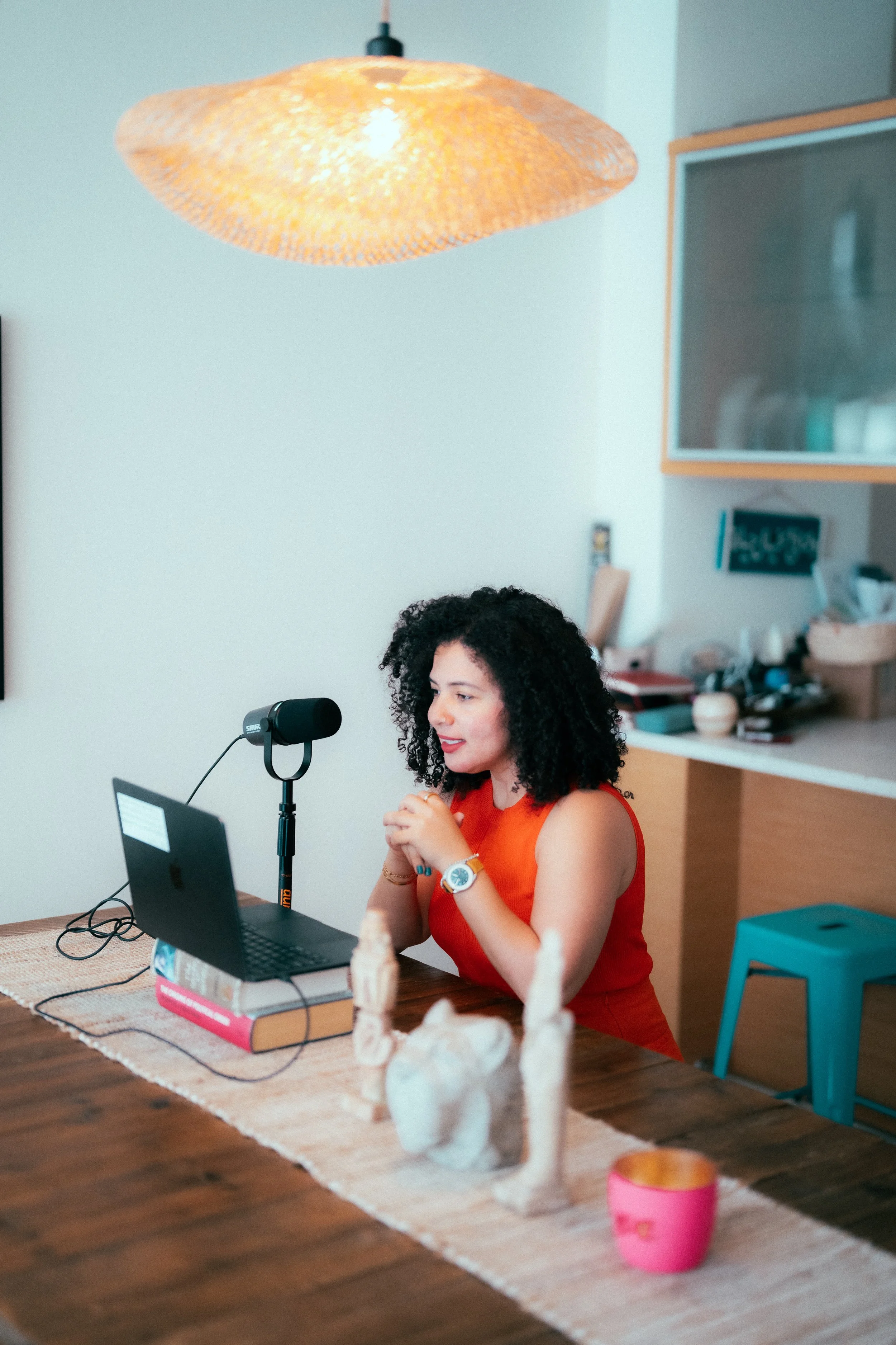 A woman with curly black hair wearing an orange sleeveless top and a white watch sitting at a wooden table with a laptop, microphone, and books, in a kitchen or dining room, with a decorative horse sculpture on the table and a ceiling light above.