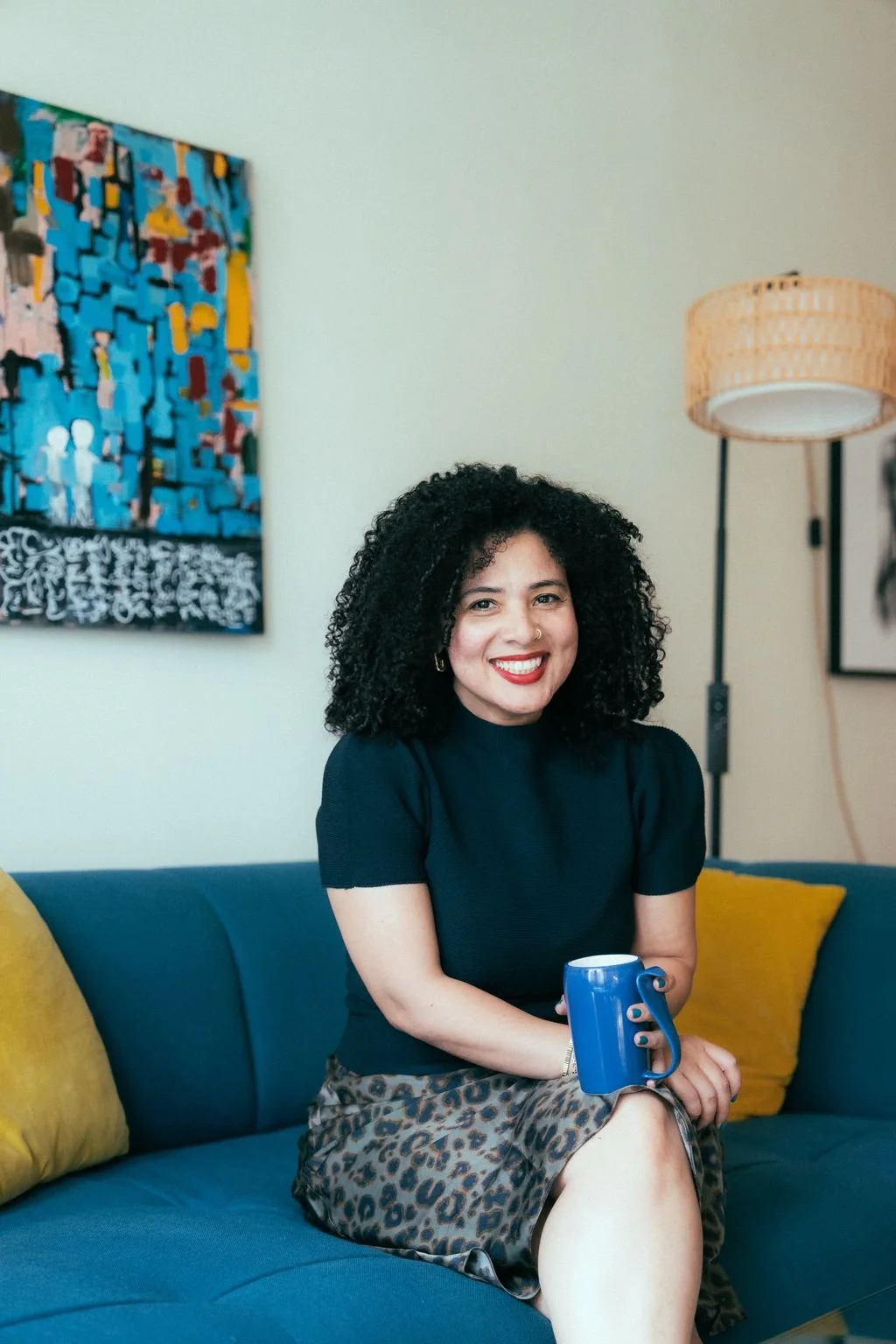 A woman with curly black hair sitting on a blue sofa holding a blue mug, wearing a black top and leopard print skirt, smiling, with colorful abstract art on the wall and a standing lamp behind her.