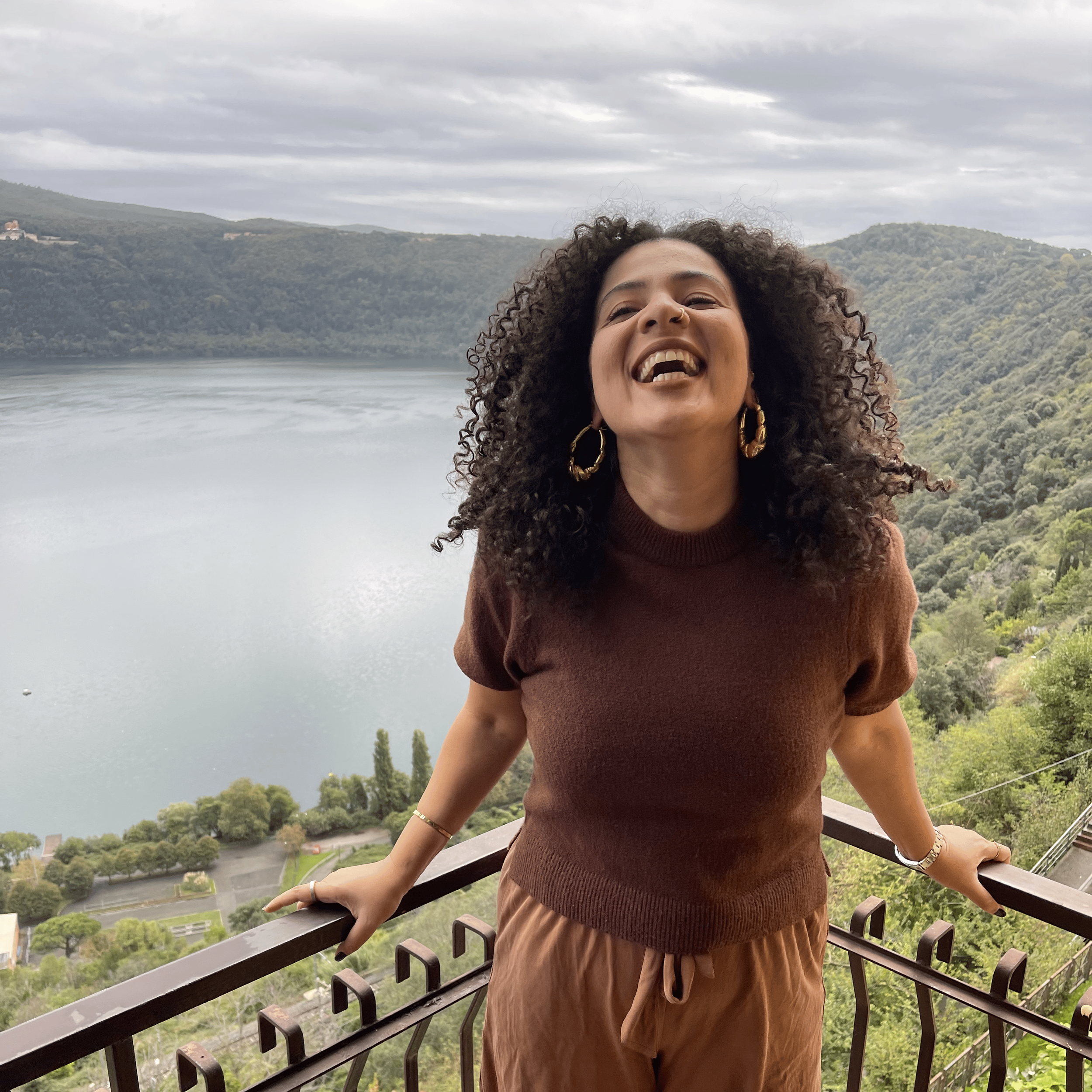 A woman with curly hair, wearing large earrings and a brown sweater, standing on a balcony with a scenic lake and mountains in the background, smiling and looking happy.