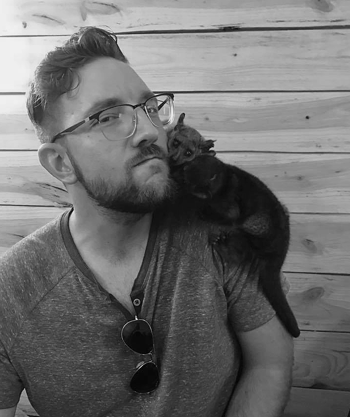 A man with glasses and a beard sitting in front of a wooden wall, with a small, dark-colored kitten resting on his shoulder.
