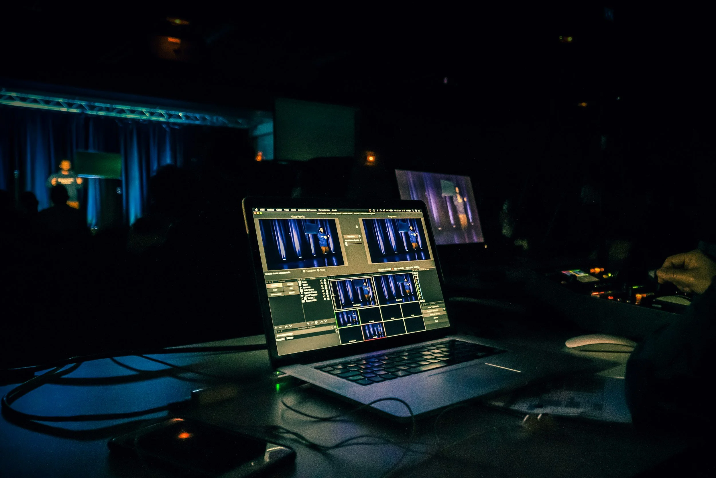 Close-up of a laptop with video editing software open, positioned on a dark table in a dimly lit environment, likely during a live event or production.