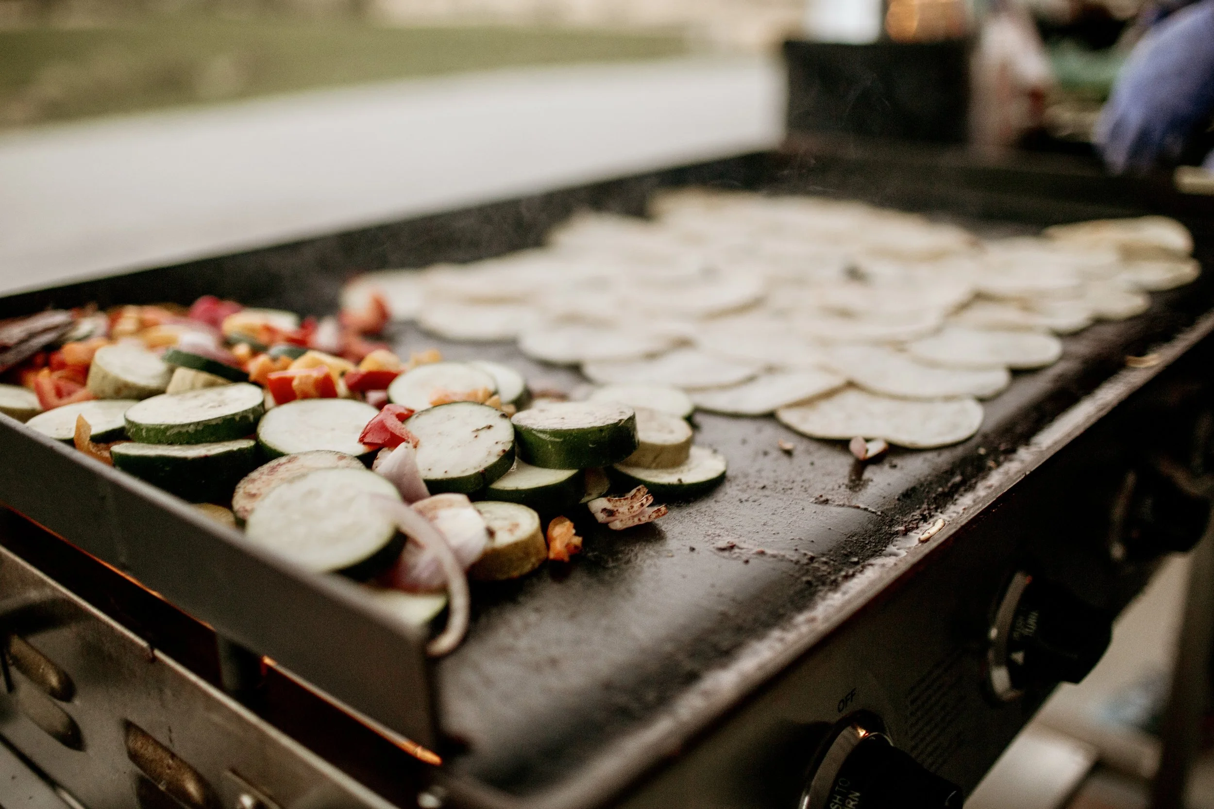 Sliced zucchini, red bell peppers, and onions on a griddle, with more sliced zucchini cooking in the background.