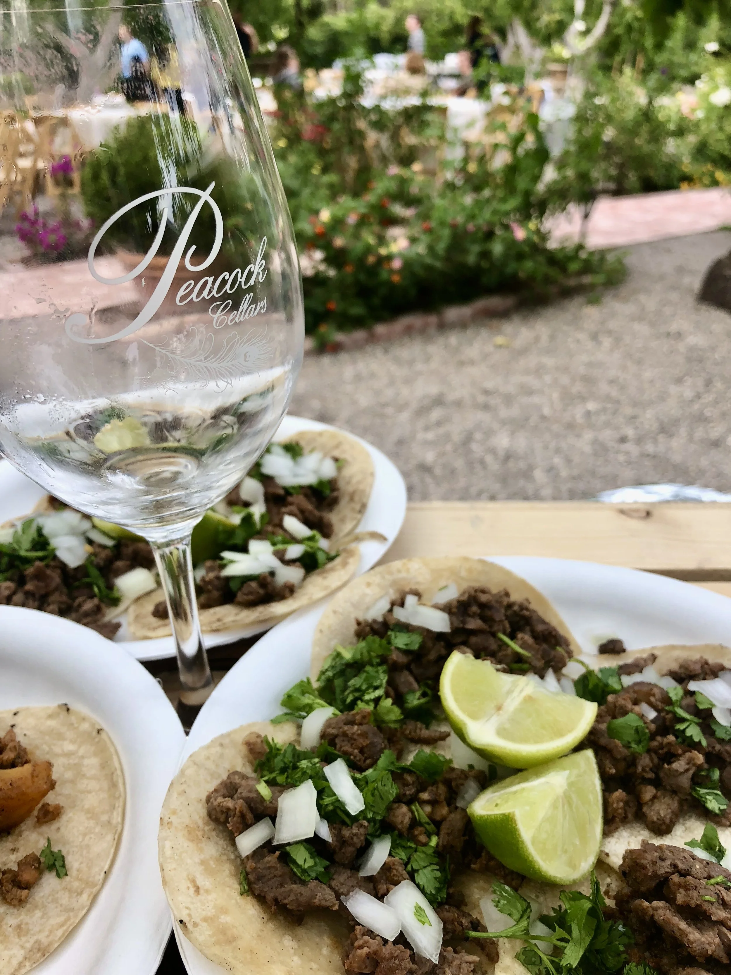 Three beef tacos topped with chopped onions and cilantro, garnished with lime wedges, on white plates. An empty Peaock Cellars wine glass is in the foreground, with a blurred outdoor garden setting in the background.
