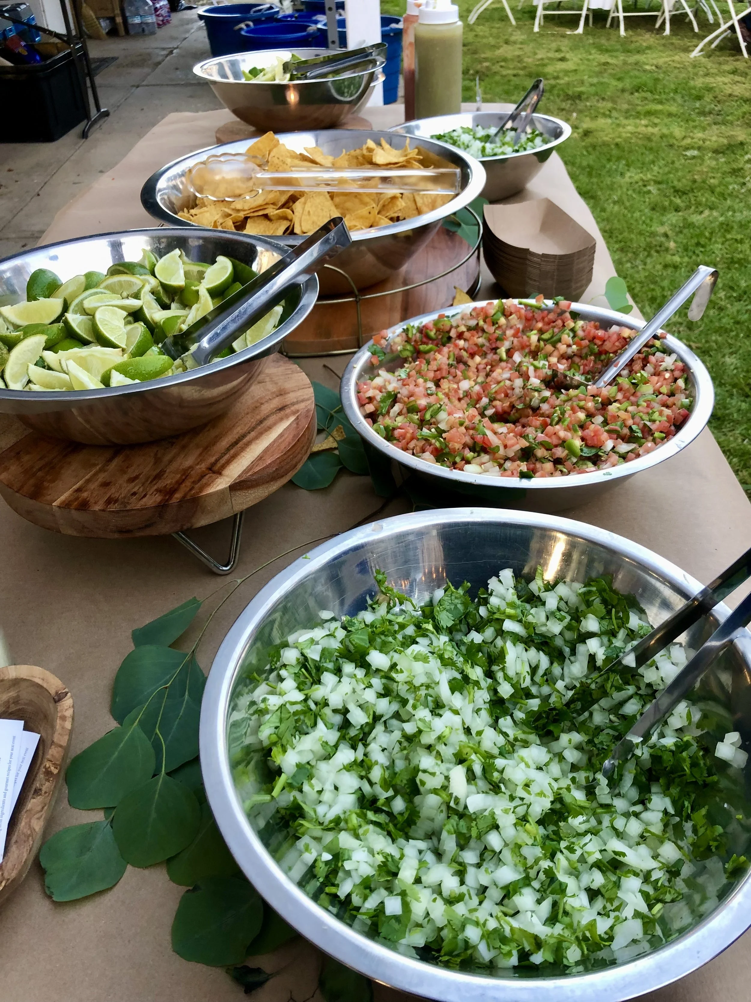 A spread of taco toppings and condiments on a table, including chopped onions and cilantro, salsa, lime wedges, shredded lettuce, tortilla chips, shredded cheese, diced tomatoes, sour cream, salsa, and condiments, set outdoors with chairs and grass i