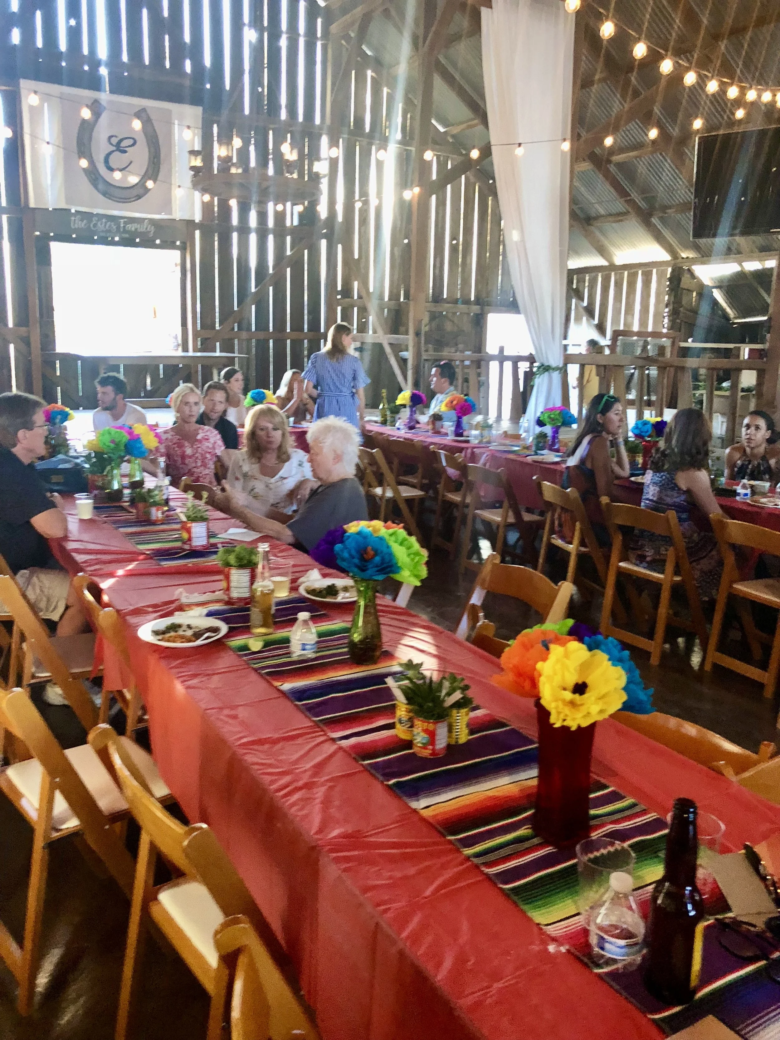 People gathered at a festive party in a rustic barn with wooden walls, decorated with colorful flowers and vibrant tablecloths.