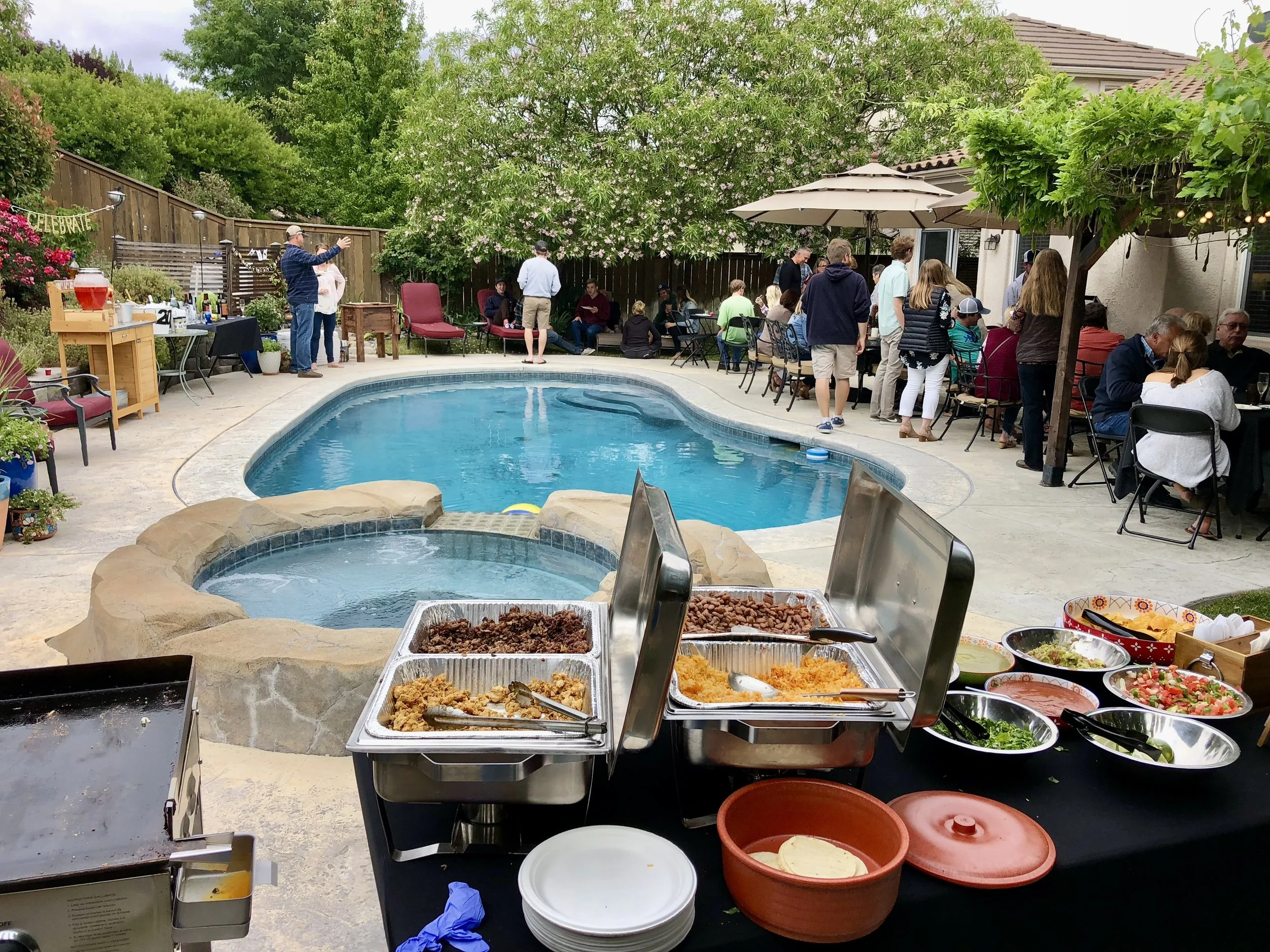 Outdoor backyard party with a swimming pool, hot tub, people gathering and dining tables under umbrellas, and buffet food setup in the foreground.