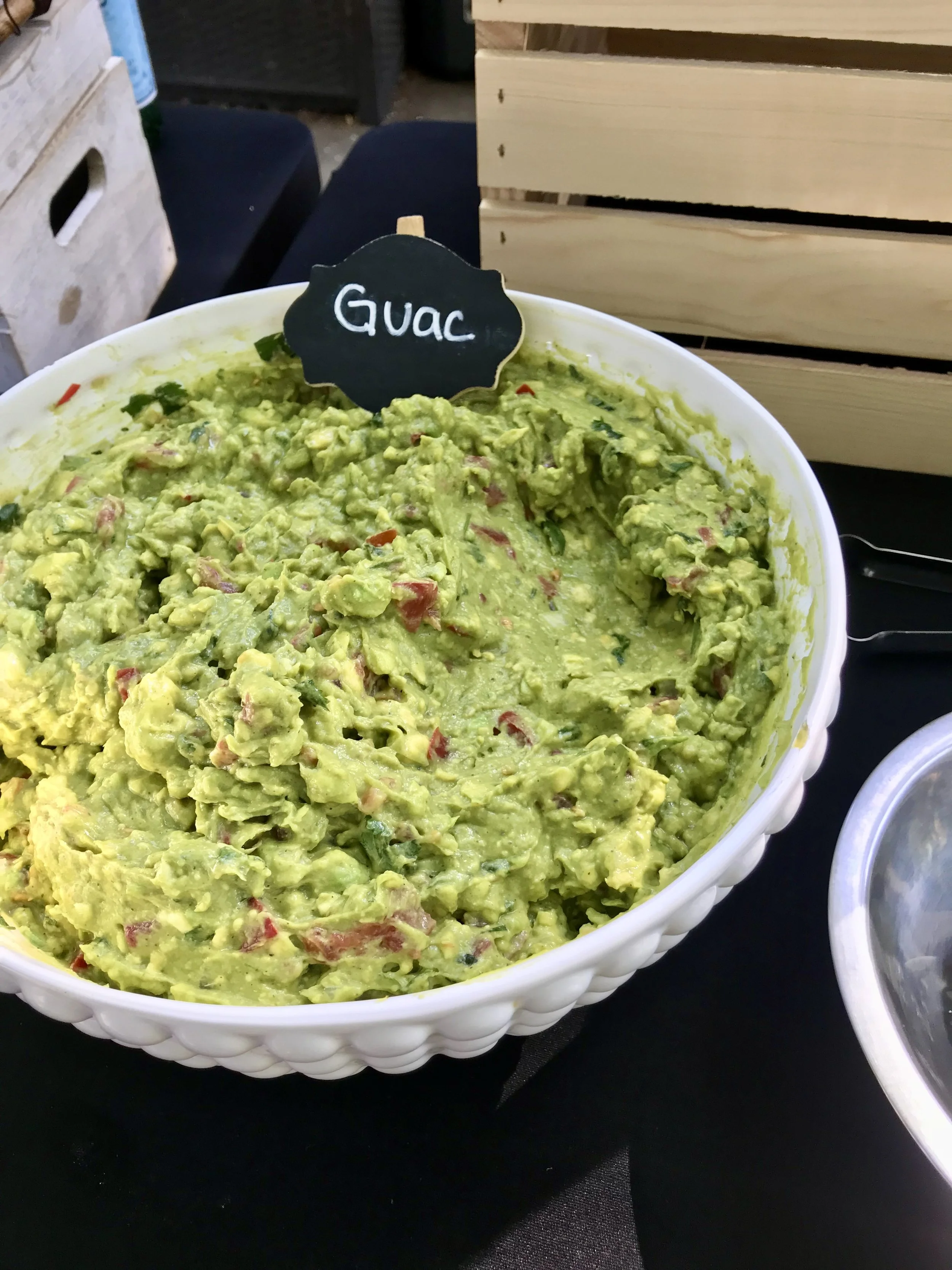 A large white bowl filled with fresh guacamole, placed on a black table. A small black chalkboard sign with the word 'Guac' written in white stands in the bowl.