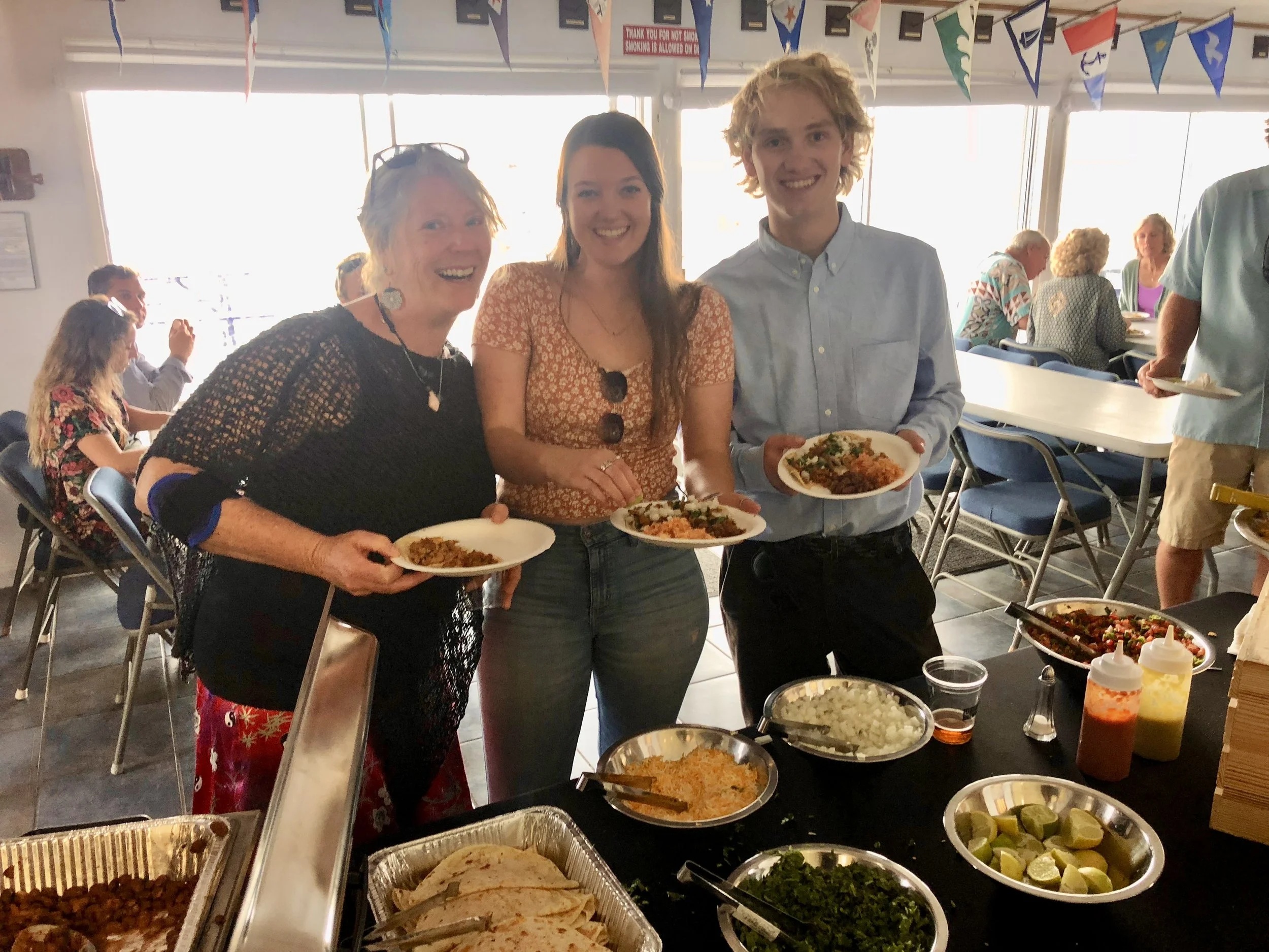 Three people smiling and holding plates of food at a buffet table during a social gathering, with other guests in the background and colorful bunting hanging overhead.