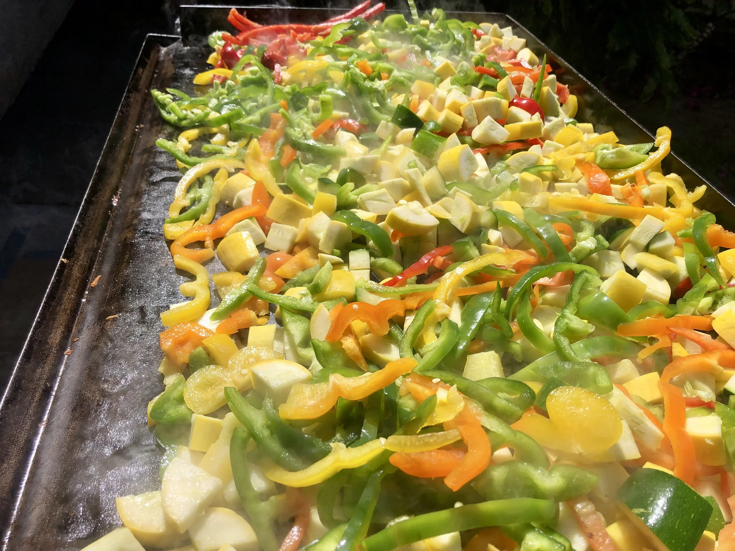 A close-up view of a large tray of grilled mixed vegetables including green, yellow, and orange bell peppers, zucchini, and red chili peppers.