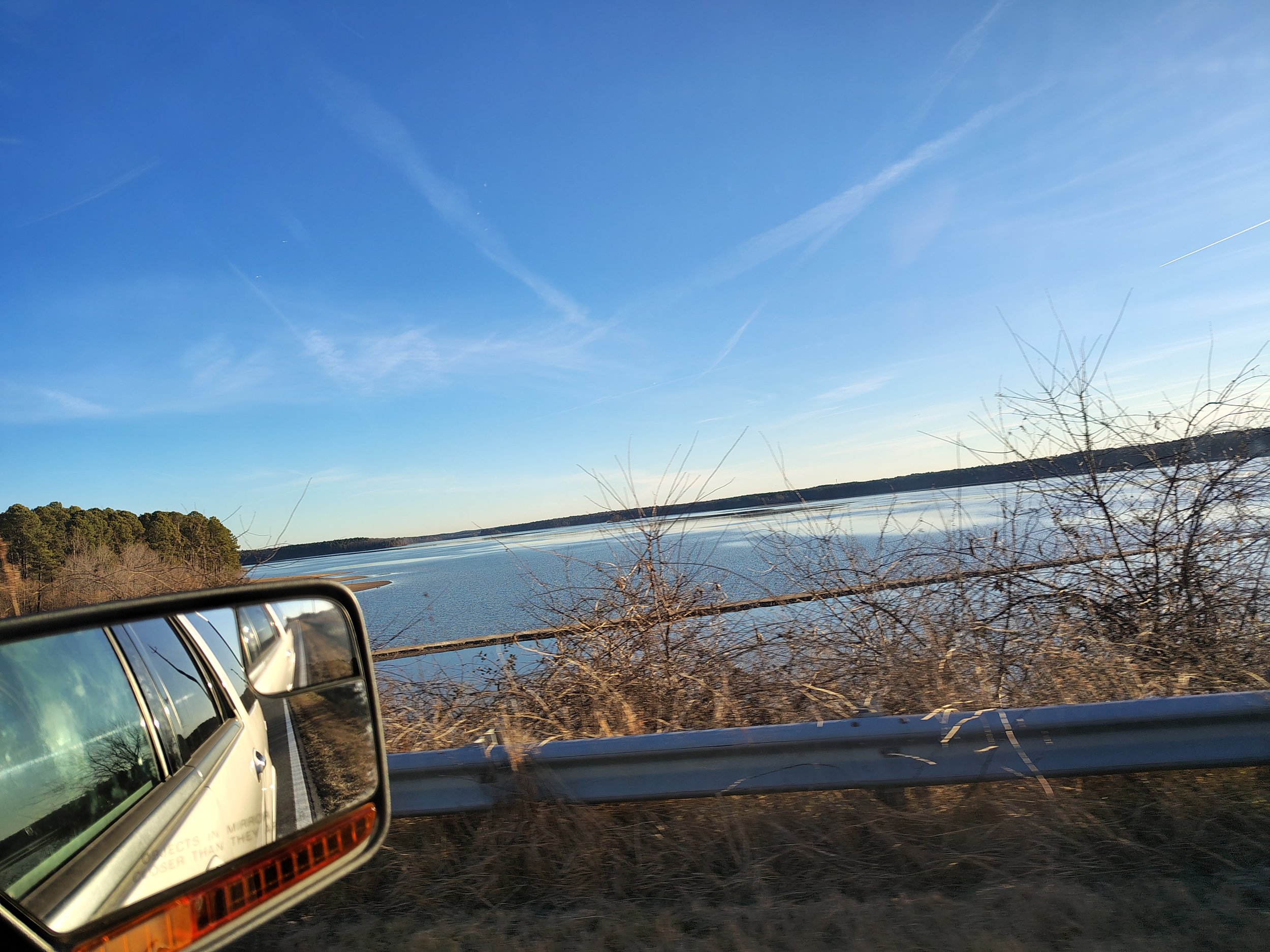 View of a body of water with a clear blue sky, trees in the distance, and dry bushes in the foreground. The photo is taken from inside a vehicle, showing part of the side mirror reflecting the road.