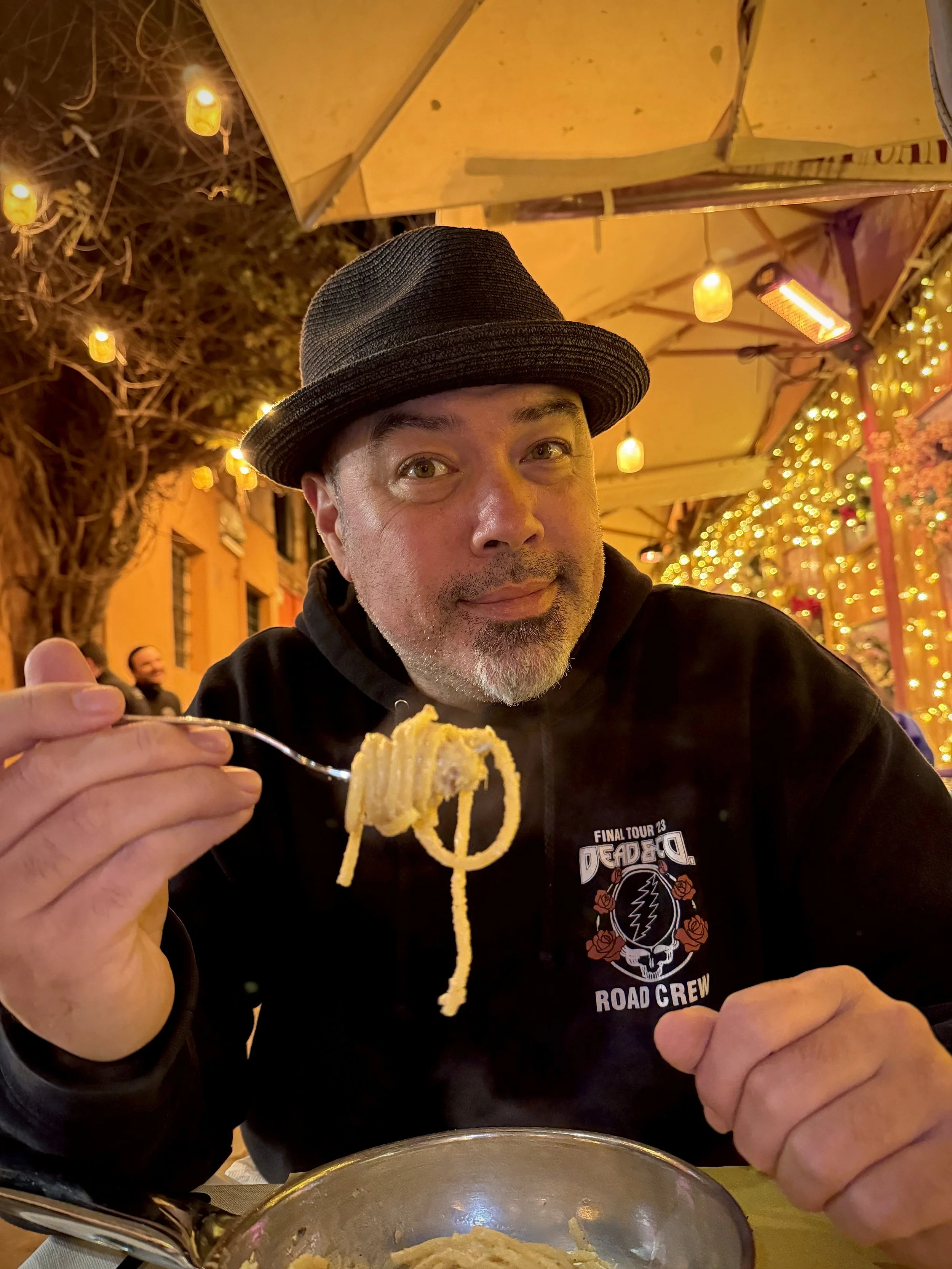 A man wearing a black fedora and a black hoodie with a 'Final Tour 23 Dead & Co Road Crew' logo is sitting at an outdoor restaurant at night. He is holding a fork with spaghetti and looking at the camera. The background is decorated with hanging yellow lights and string lights, creating a cozy atmosphere.
