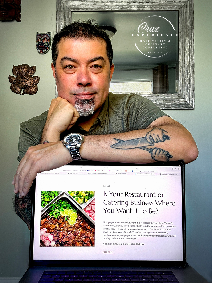 Man with tattoos and a watch smiling and resting his chin on his hand, sitting behind a laptop displaying an article about restaurant and catering business, with a mirror and decorative wall art in the background.
