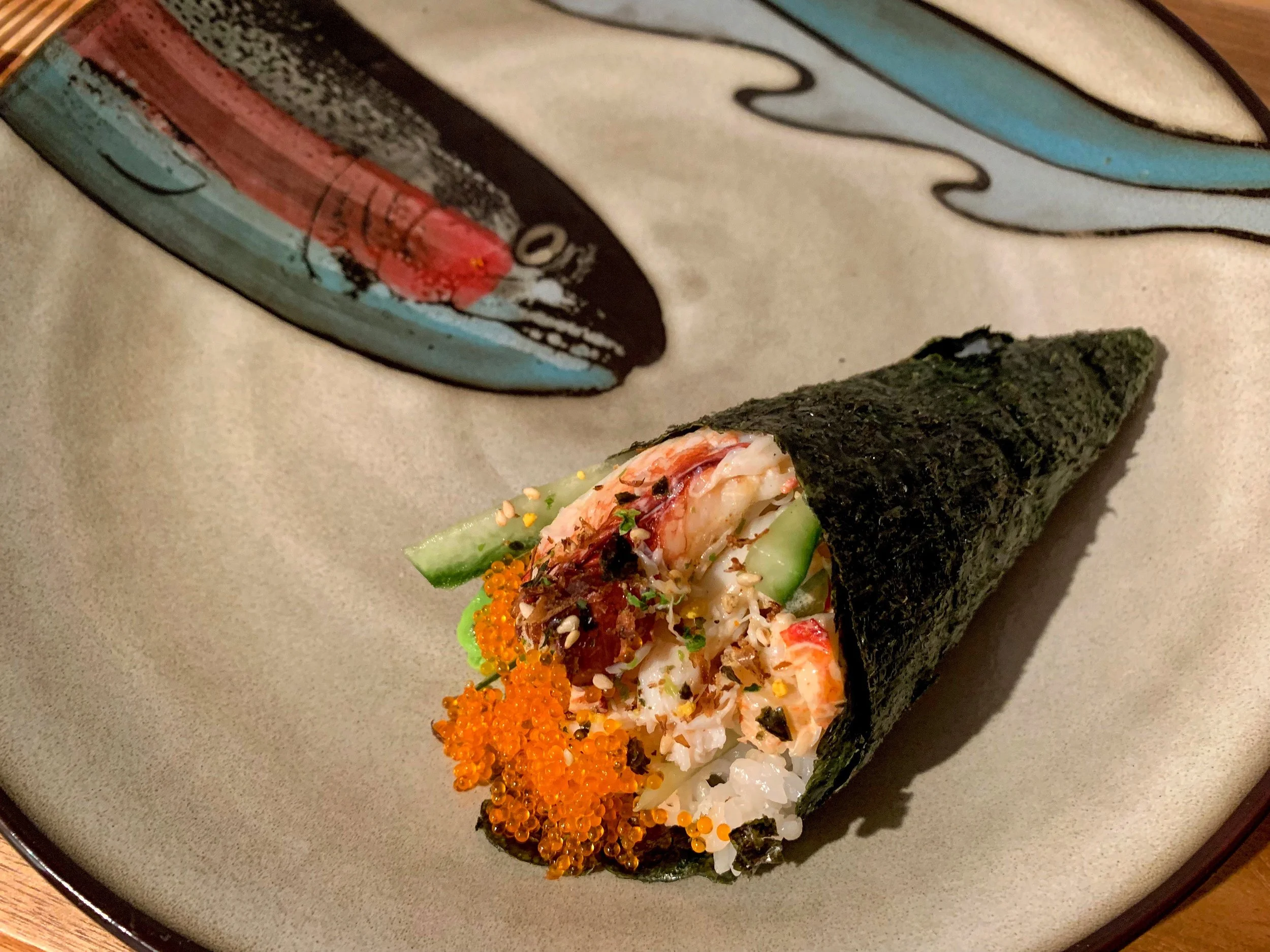 A sushi hand roll with nori seaweed wrapping, filled with rice, shrimp, cucumber, fish roe, and other ingredients, placed on a beige plate with a decorative design.