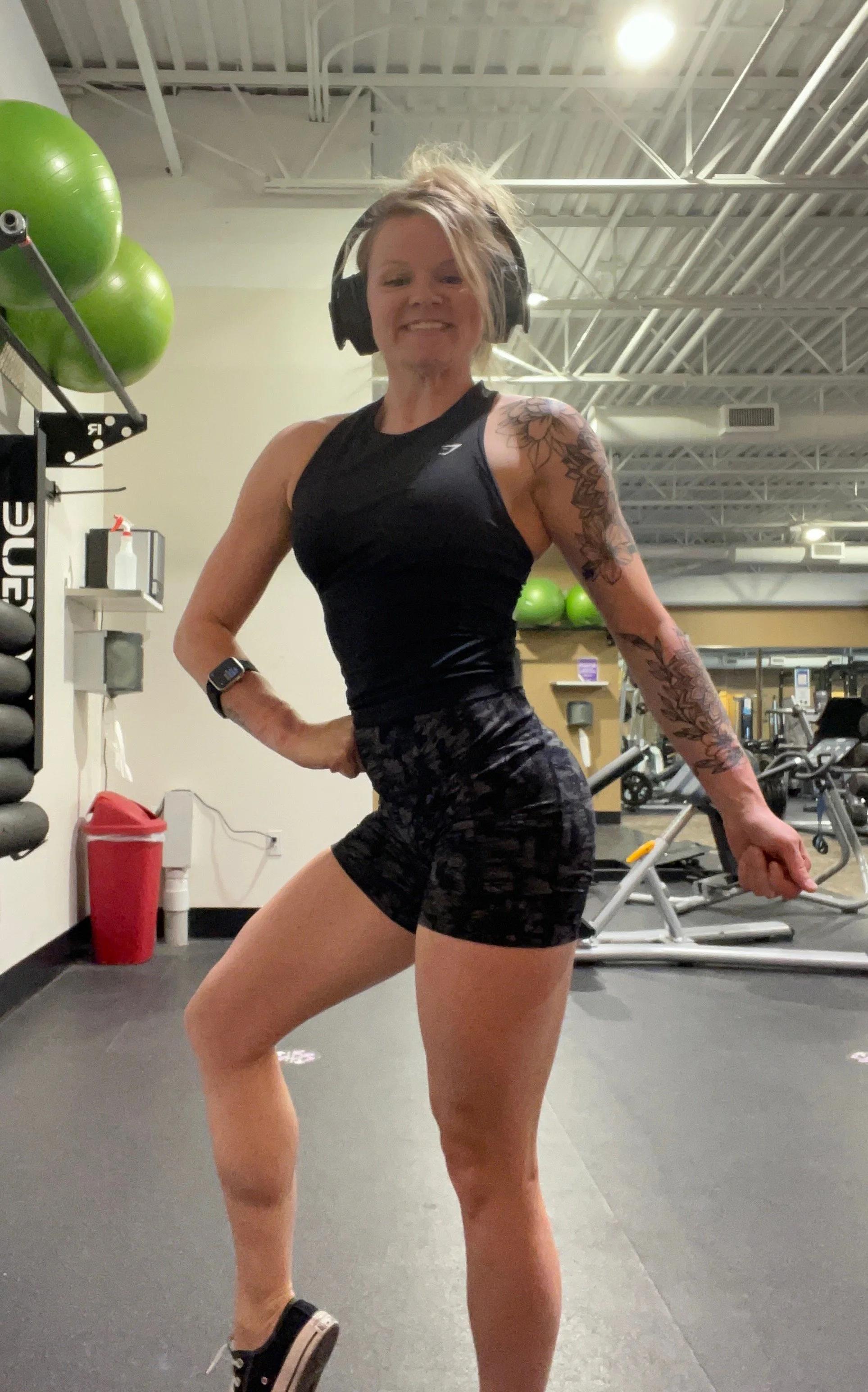 A woman in workout attire posing confidently at the gym, wearing headphones, a black tank top, and camo-patterned shorts, with tattoos on her arm.