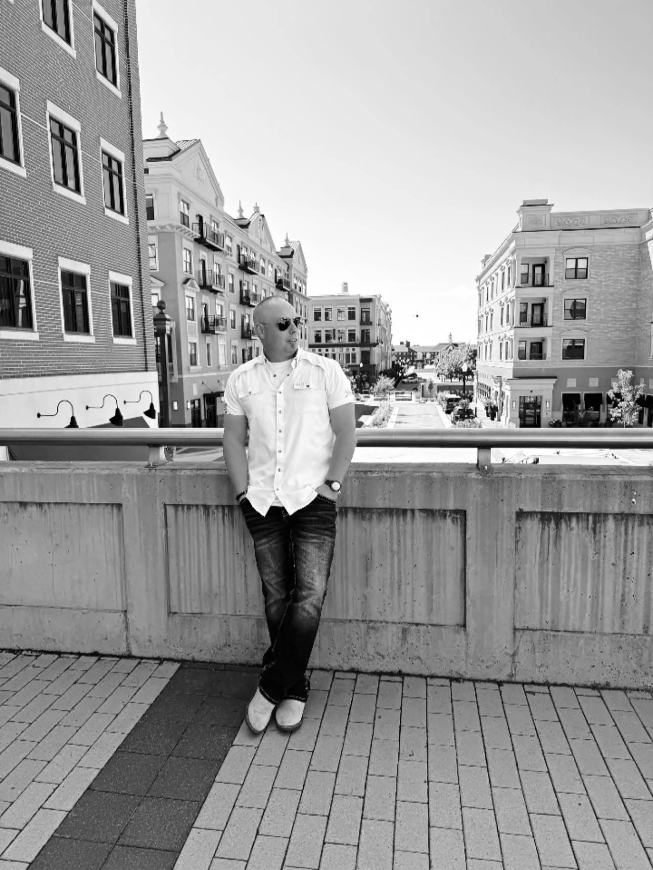 A man wearing sunglasses, a short-sleeved shirt, and jeans is leaning against a concrete ledge on a city street in black and white. His hands are in his pockets, and he looks to the side. Behind him are multi-story buildings with balconies and lamp posts.