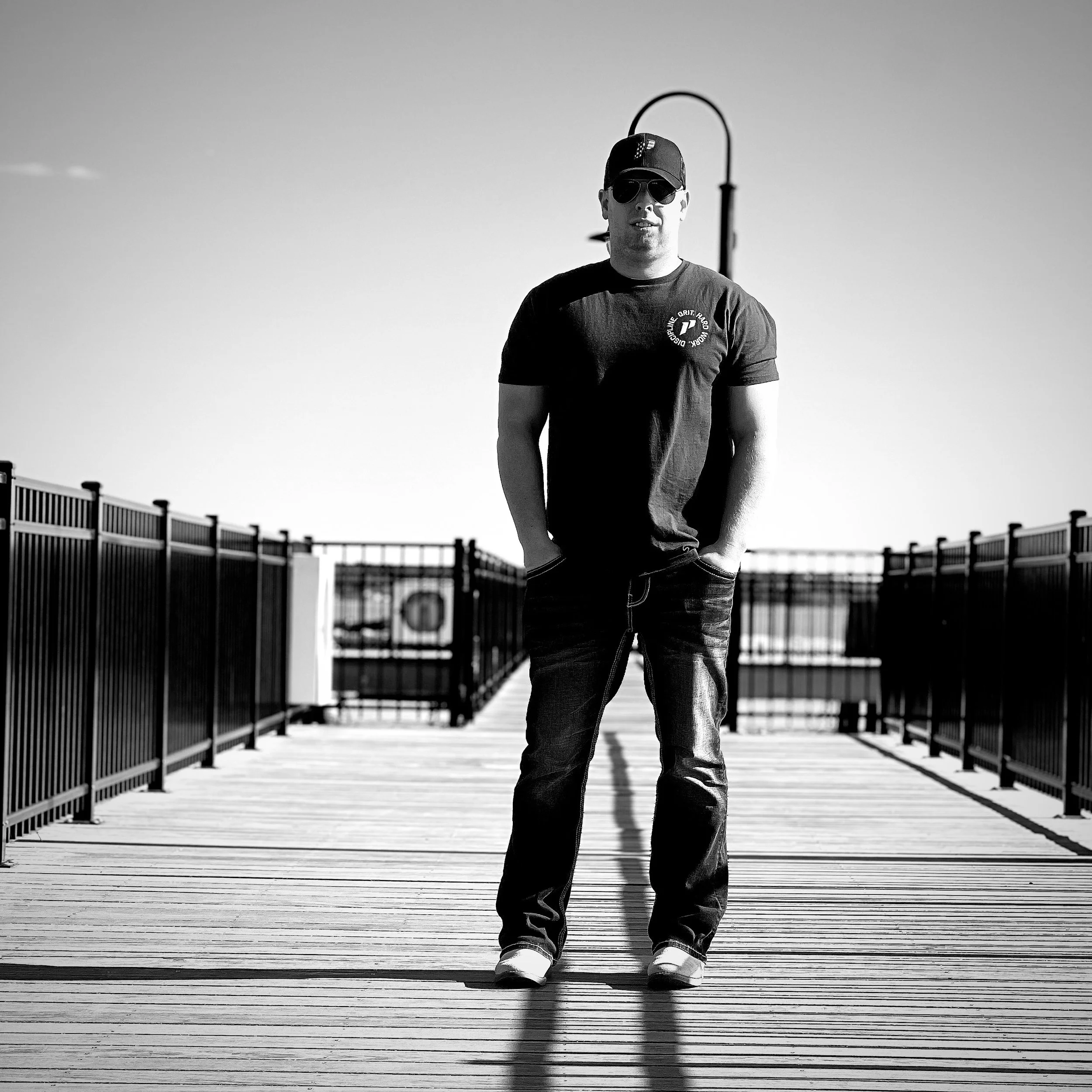 A man wearing sunglasses, a baseball cap, a dark t-shirt, and jeans standing on a wooden pier with handrails, with a lamp post behind him, in black and white.