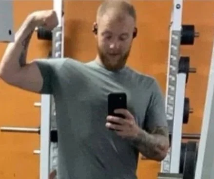 A young man with a beard taking a selfie in a gym while flexing his bicep, with gym equipment in the background.