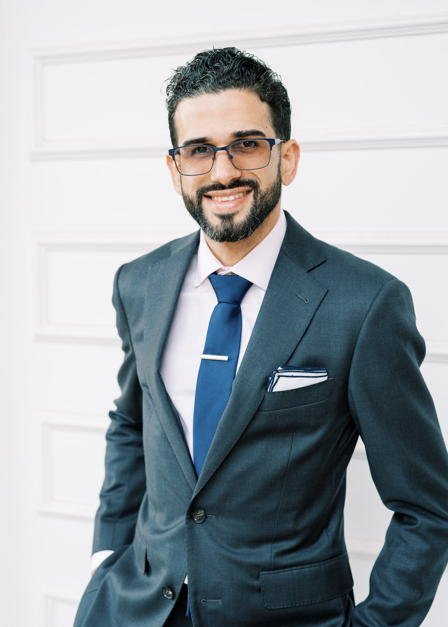 A man in a dark gray suit, white shirt, and blue tie with a silver tie clip, smiling and standing in front of a white wall with paneling.