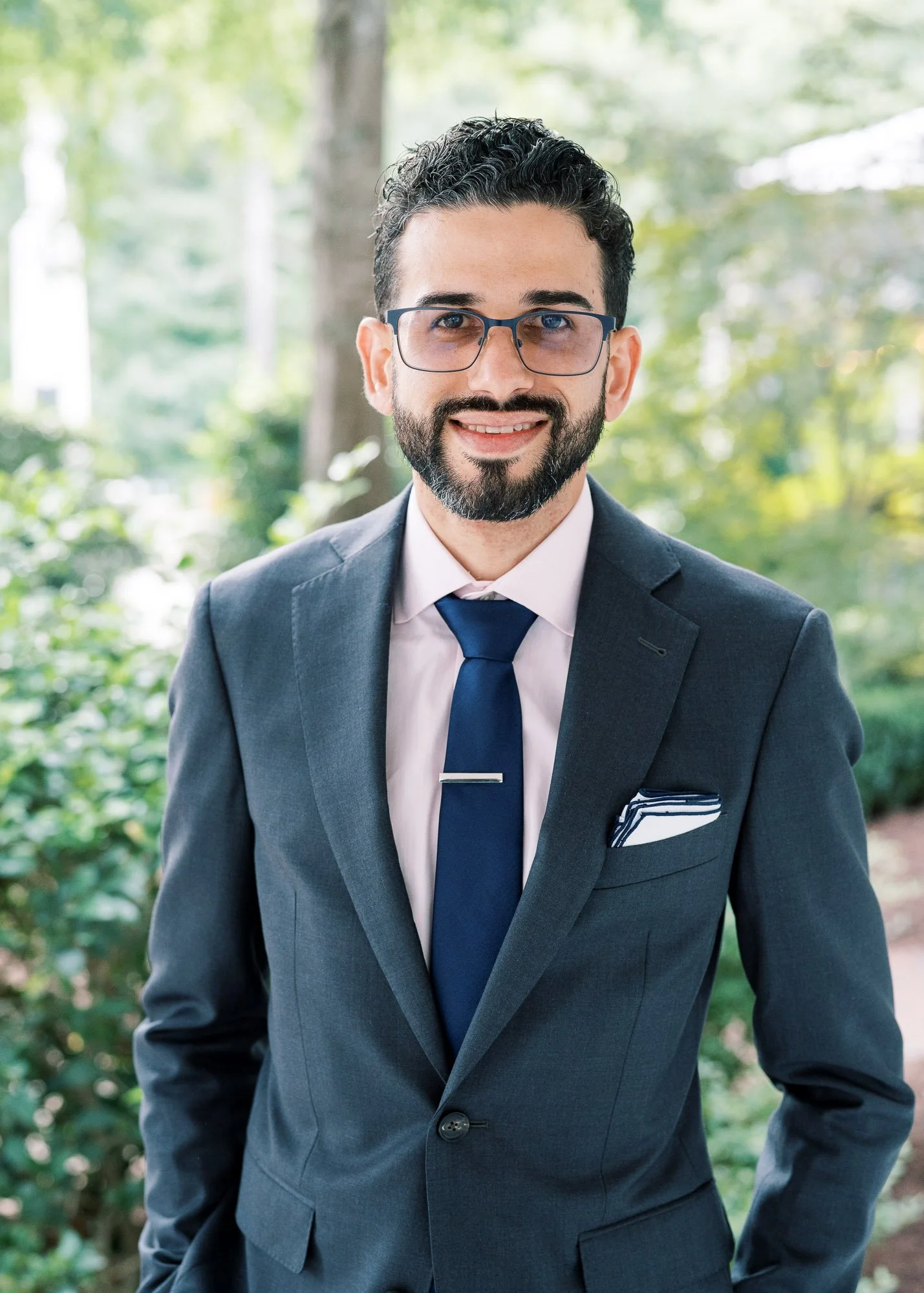 A man with dark curly hair, a beard, and glasses wearing a navy blue suit, light pink shirt, and navy tie, smiling outdoors with greenery in the background.