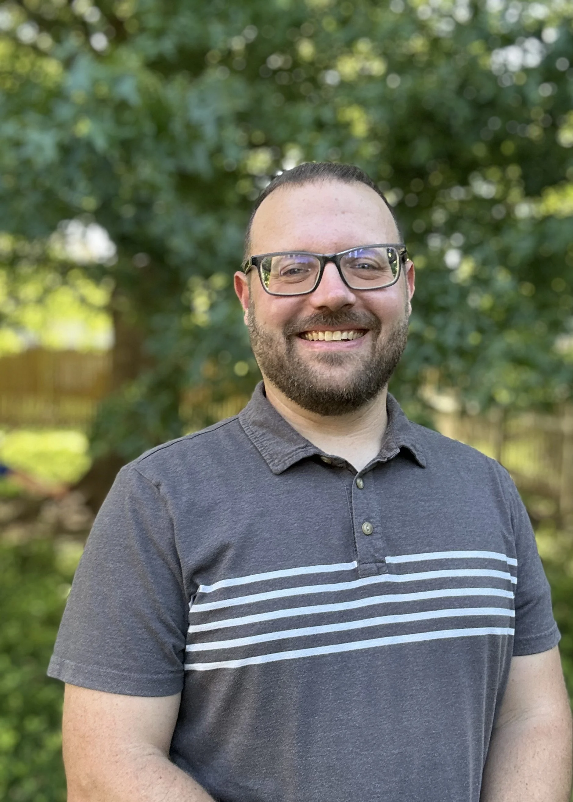 A smiling man with glasses and a beard, wearing a gray polo shirt with white horizontal stripes, standing outdoors with a background of green trees and sunlight.