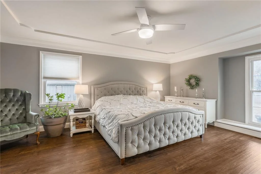 A bedroom with a large tufted bed, two nightstands with lamps, a gray armchair, a potted plant, and a white dresser with decorative items and a wreath, wooden floor, and two windows with blinds.
