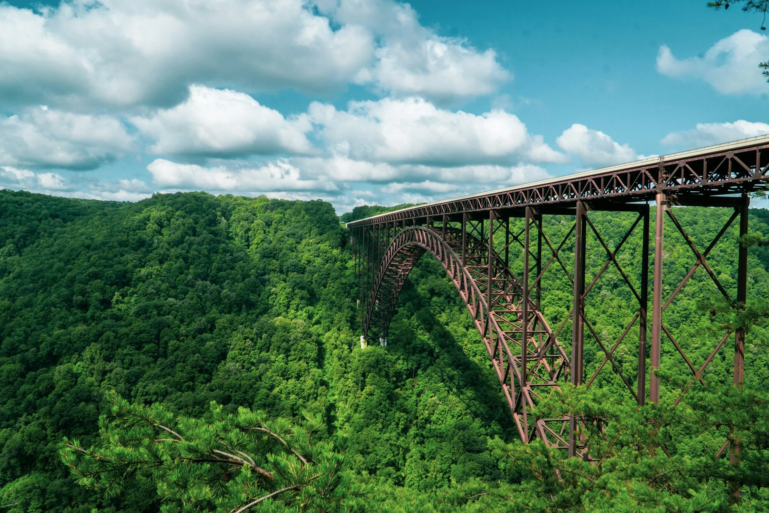 A large steel arched bridge extending over a green valley with trees and hills under a blue sky with white clouds.