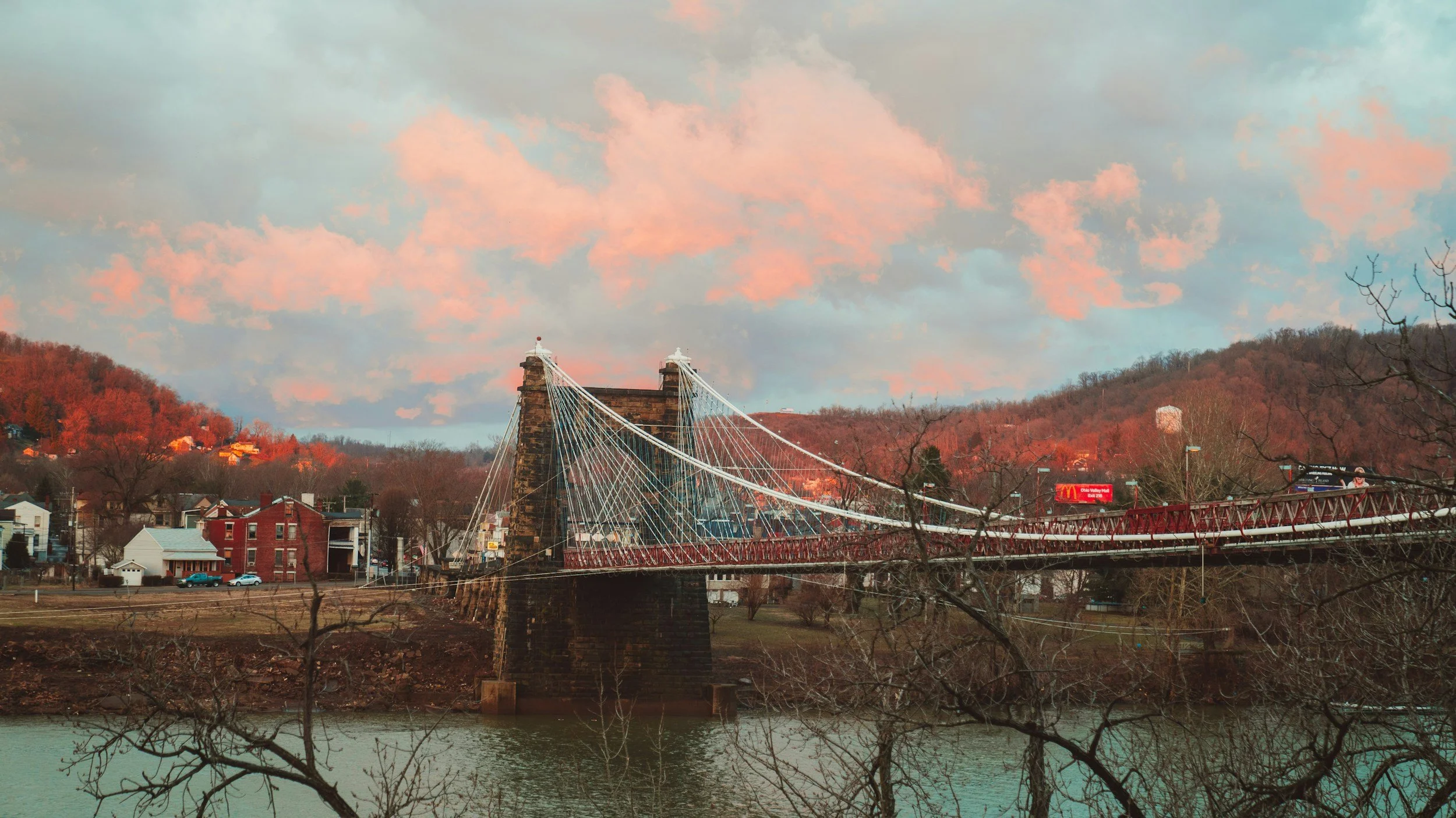 A suspension bridge spanning a river, with colorful houses and trees on hills in the background, under a sky with pink and gray clouds.