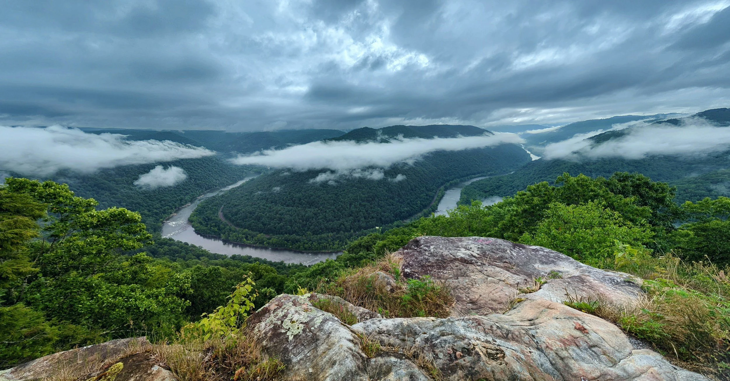 A scenic view of a winding river through dense green forested mountains under a cloudy sky, with clouds obscuring some mountain tops, taken from a rocky overlook.