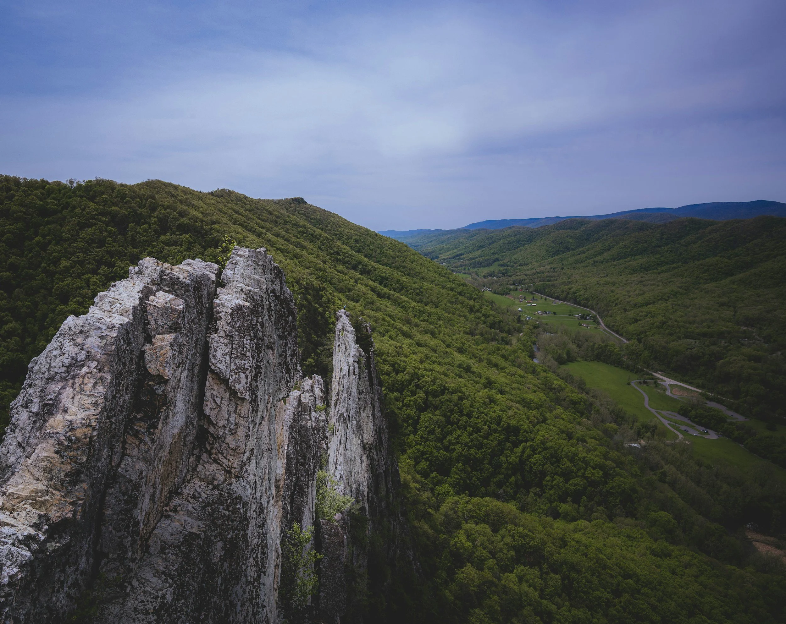 Rock formations on a mountain with a green valley and winding road below, and a cloudy sky above.