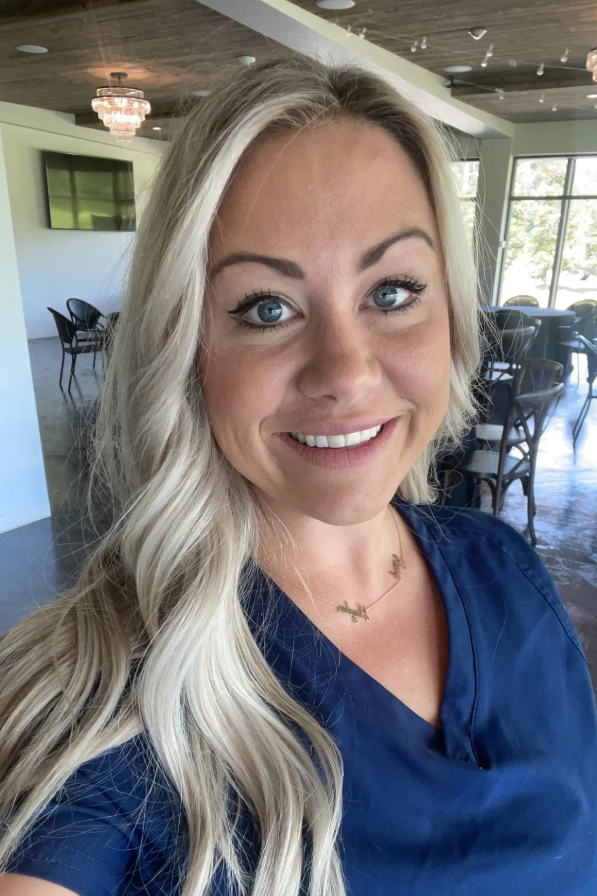 A woman with long blonde hair, blue eyes, and a bright smile, taking a selfie inside a well-lit room with large windows, a TV, and a chandelier.