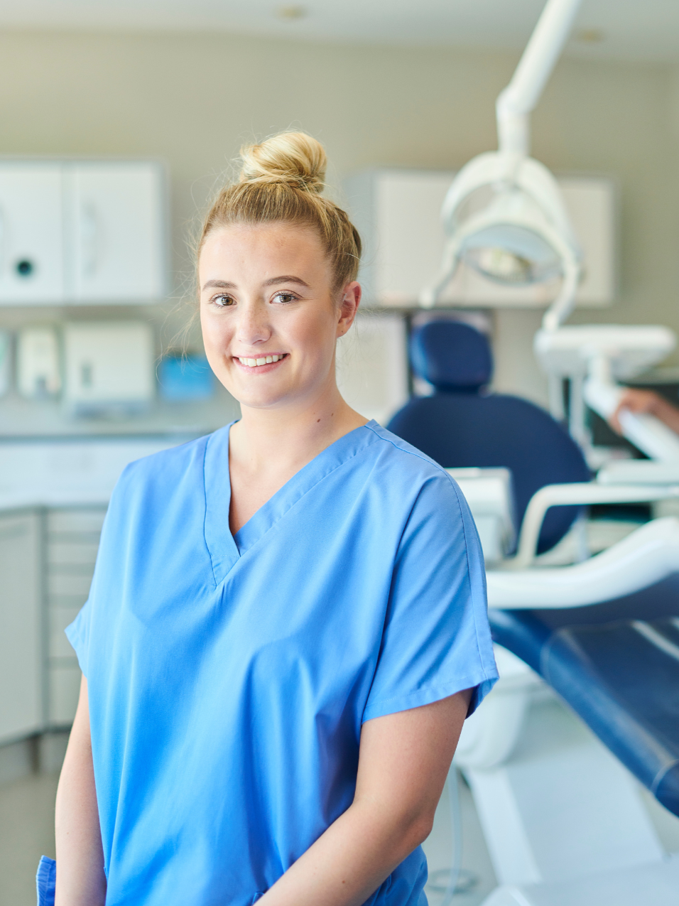 Young female dental professional in blue scrubs standing in a dental clinic with dental equipment and a chair in the background.