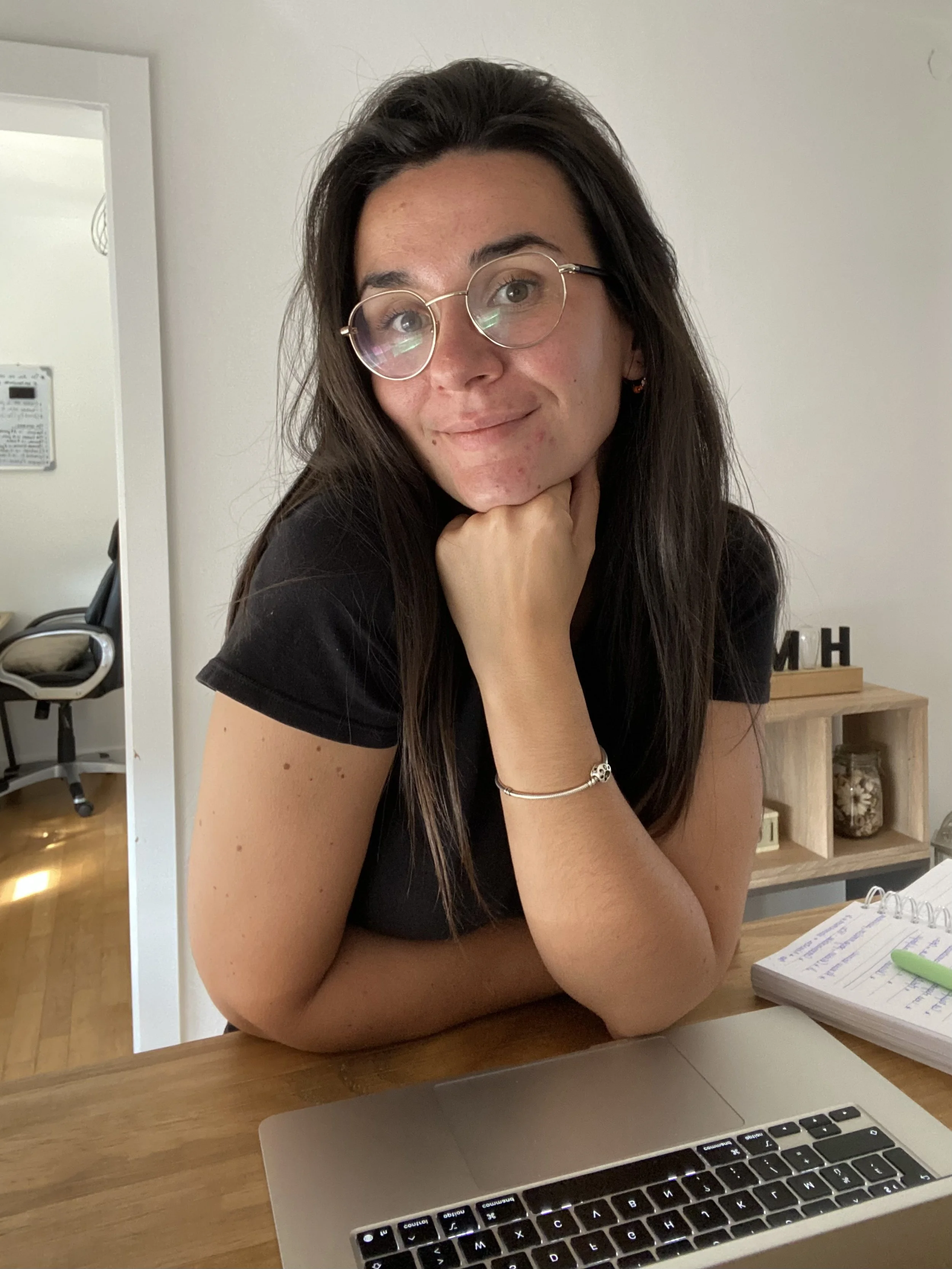 Une femme à lunettes assise à un bureau, souriant, avec un ordinateur portable, un carnet et un stylo à côté.