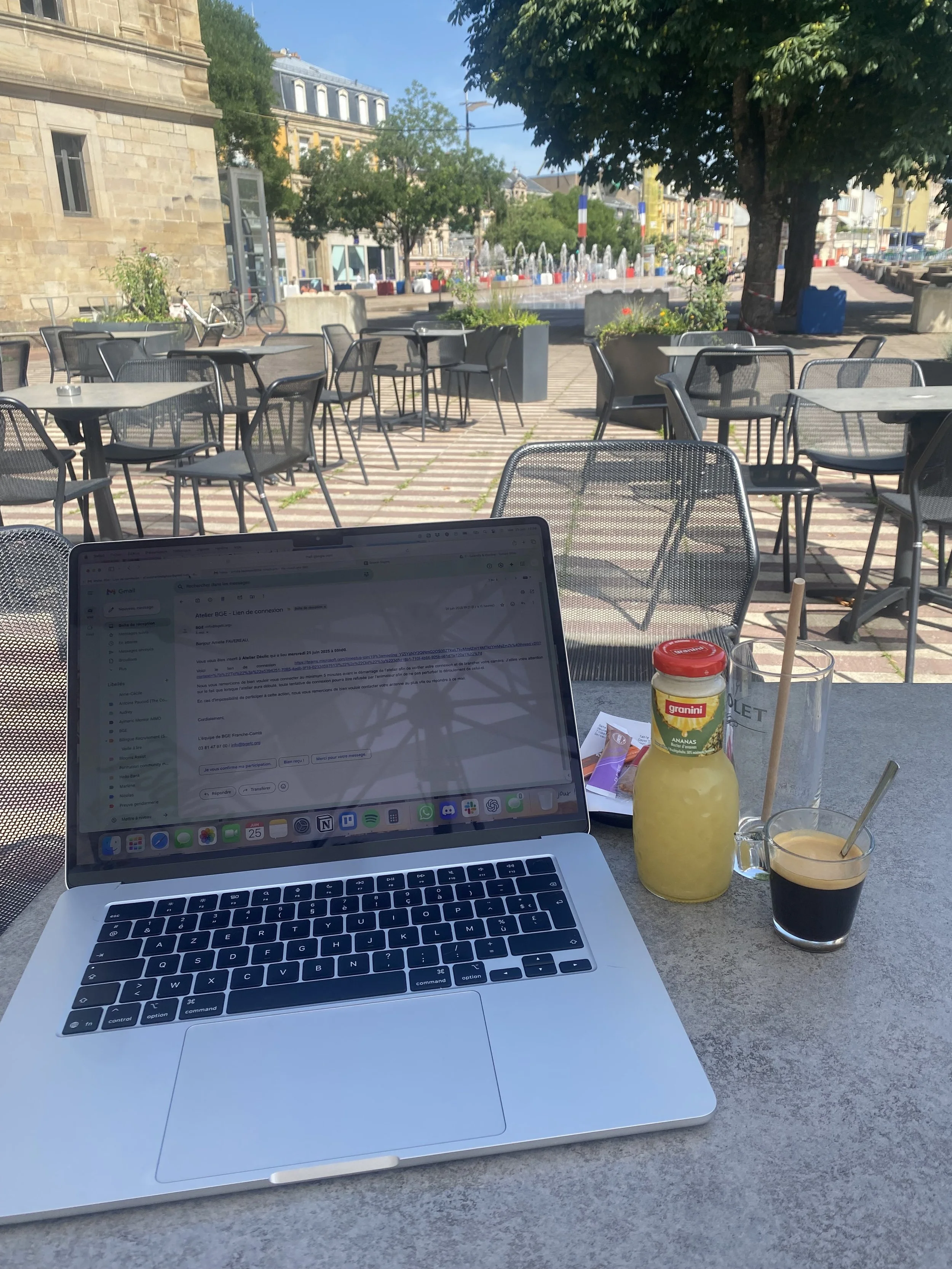 Table en extérieur avec un ordinateur portable, une bouteille de jus, un verre d'eau, un verre de café, et des papiers, avec des chaises et une fontaine en arrière-plan dans un espace public en plein air par temps ensoleillé.