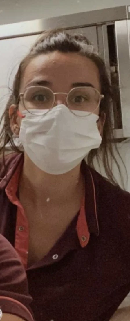 Une femme portant des lunettes, un masque facial et une chemise rouge foncé, assise dans une pièce intérieure.